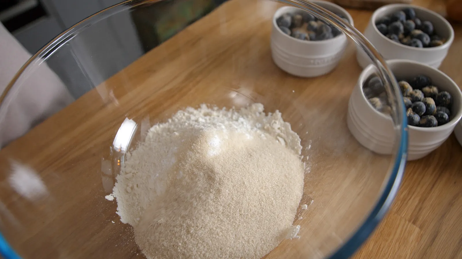 A close-up of a large clear glass bowl containing white flour and light brown sugar, with several small white ramekins filled with blueberries in the background.