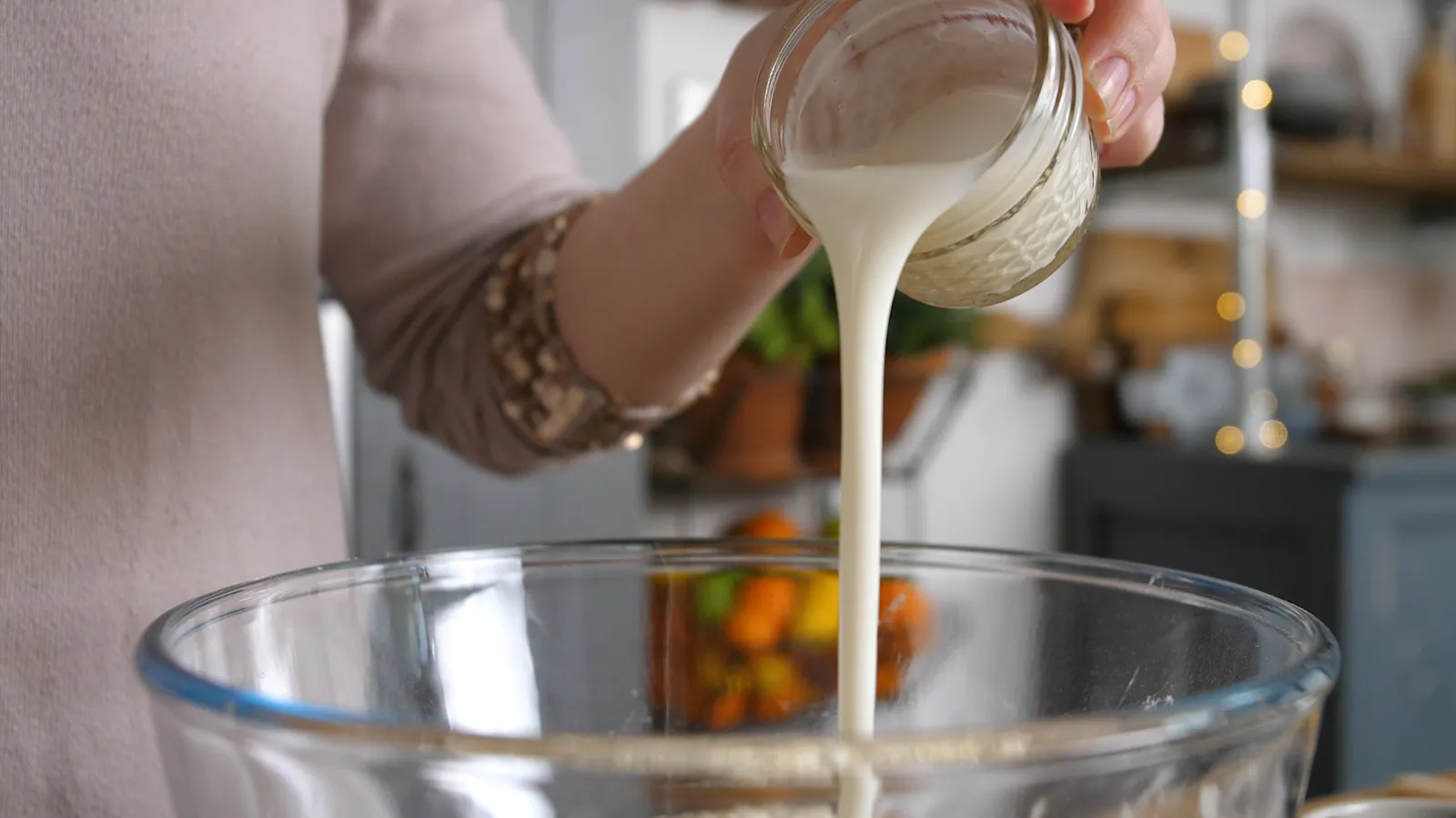 A hand pours milk from a small glass jar into a large clear glass bowl containing dry ingredients.
