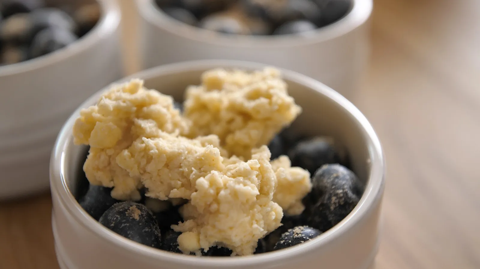 A close-up of a white ramekin filled with fresh blueberries topped with a lumpy, light-colored dough, ready for baking.