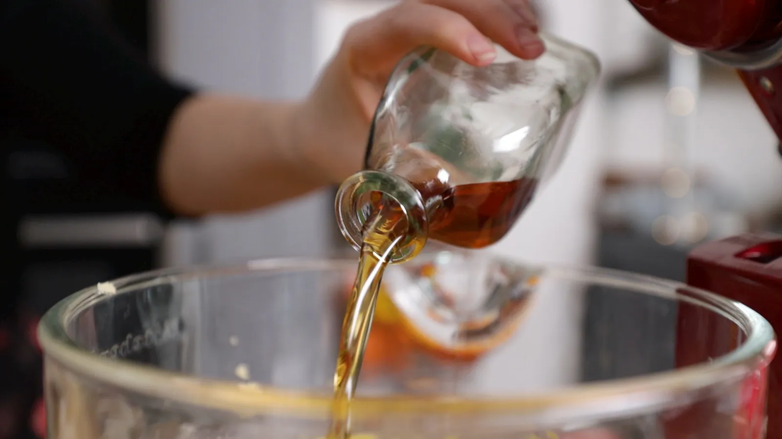A hand pours a stream of amber liquid (likely syrup or oil) from a small glass bottle into a large clear glass bowl.
