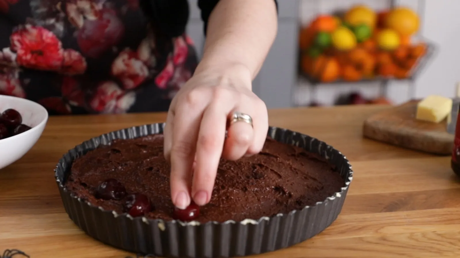 A hand places fresh red cherries onto a dark chocolate tart batter in a fluted tart pan.