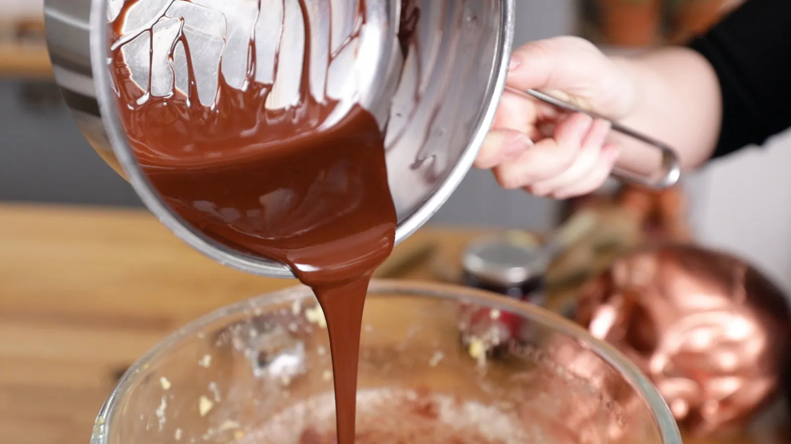 A hand pours melted dark chocolate from a stainless steel saucepan into a large clear glass bowl.
