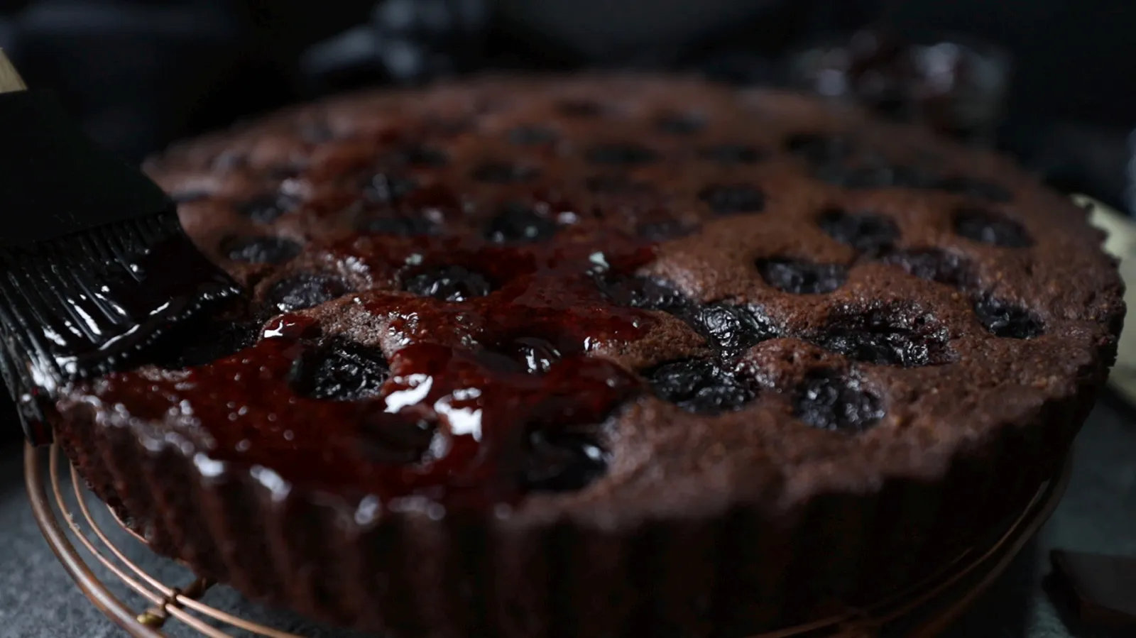 A hand uses a black pastry brush to glaze a baked chocolate and cherry tart with a shiny red sauce on a wire rack.