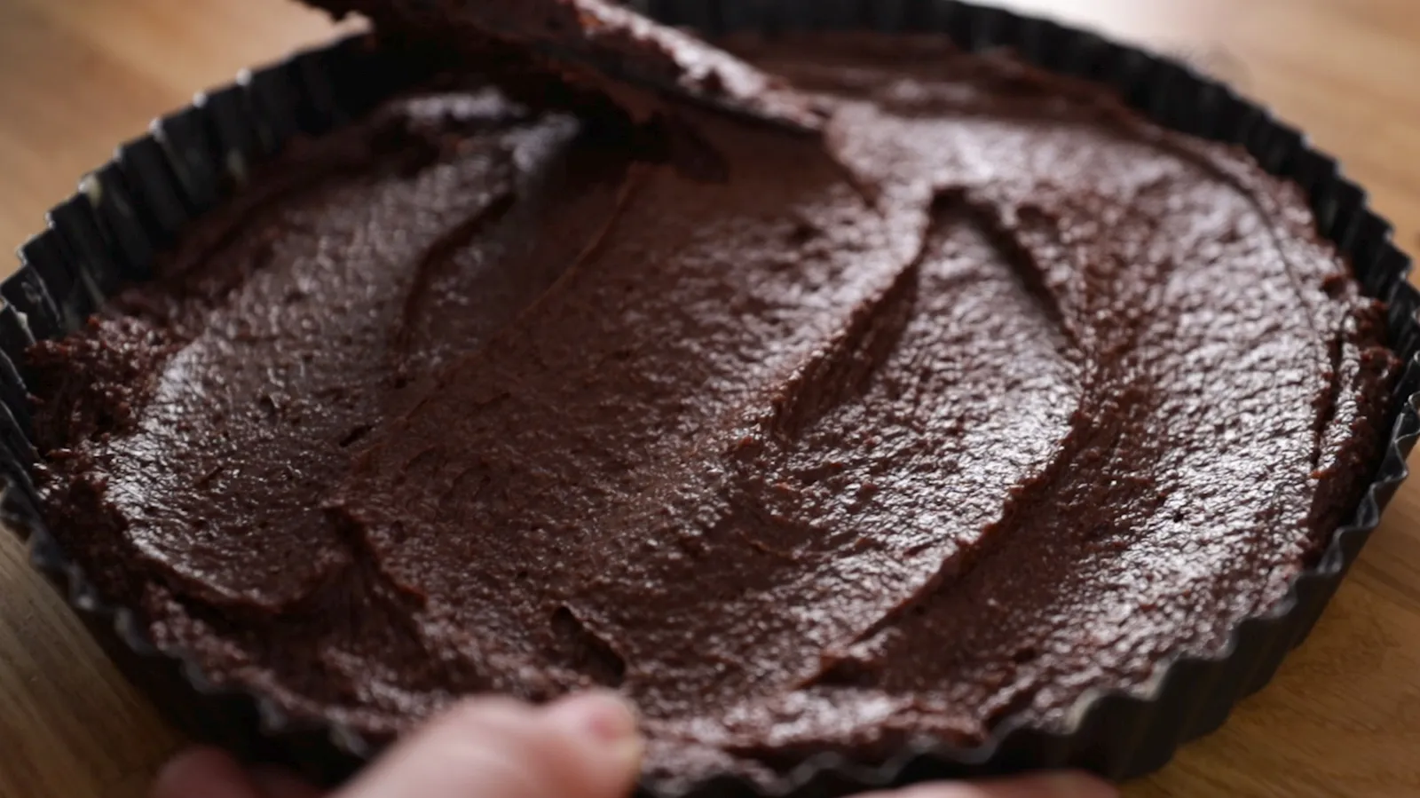 A hand uses a spatula to spread a thick, dark chocolate batter evenly into a fluted tart pan.