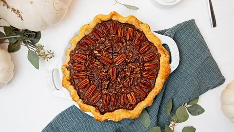 Top-down view of a freshly baked pecan pie with a golden crust on a green cloth, surrounded by white pumpkins and eucalyptus leaves.