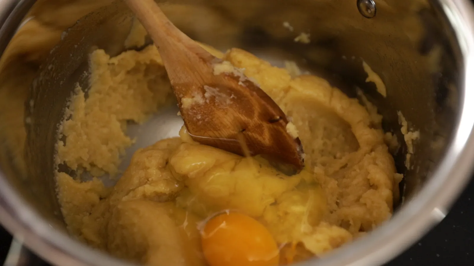 A wooden spoon mixes a light brown choux pastry dough in a stainless steel saucepan, with a cracked egg yolk visible in the mixture.