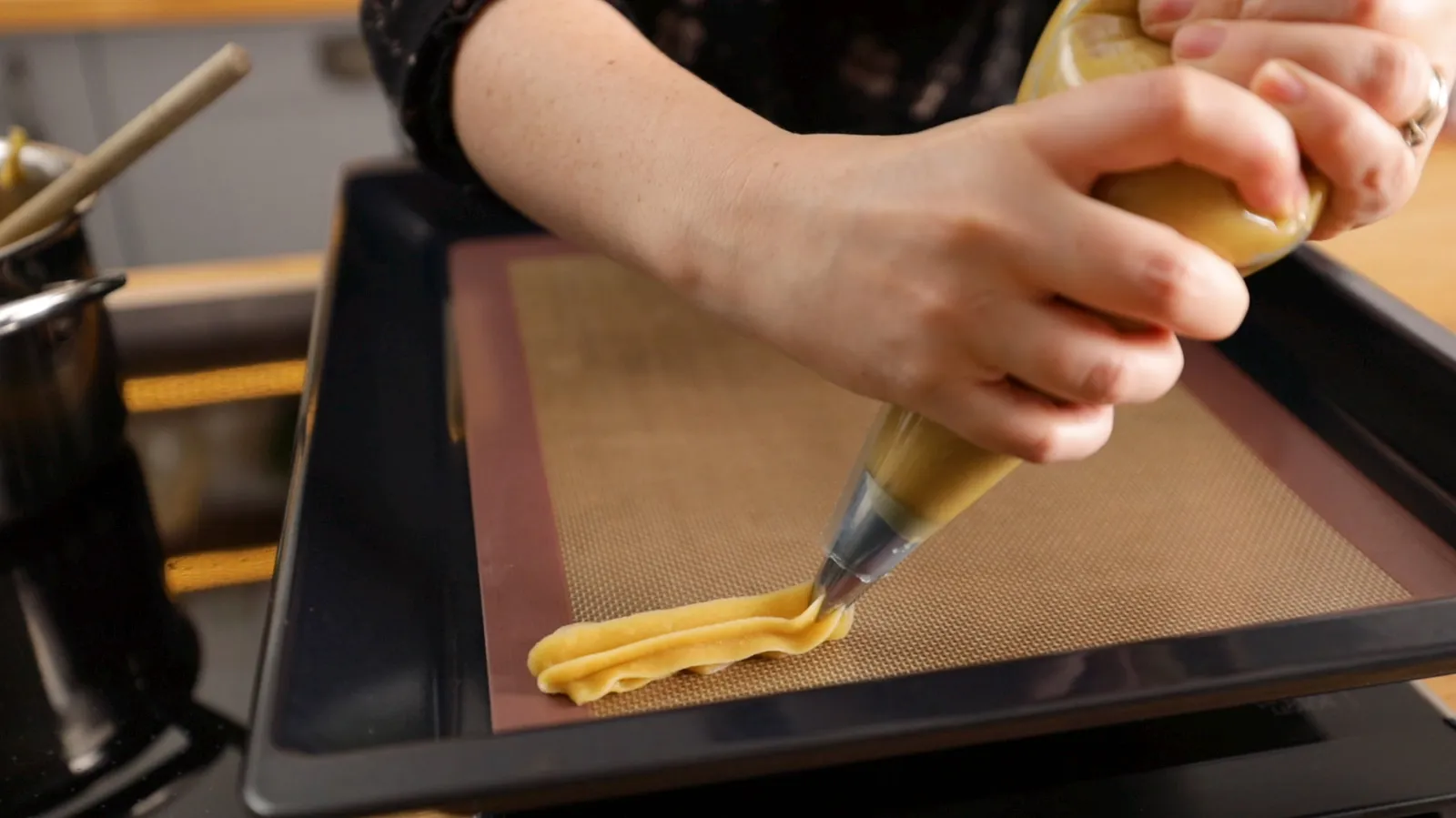 Hands pipe long, ridged strips of light brown choux pastry onto a baking tray lined with a silicone mat.