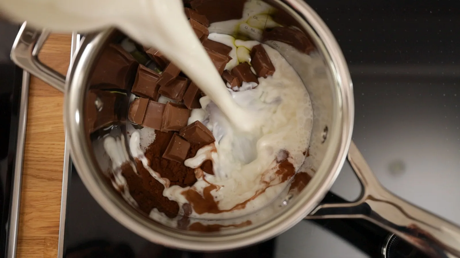 An overhead view of a stainless steel saucepan on an induction cooktop, containing dark chocolate pieces, cocoa powder, and white milk being poured in.