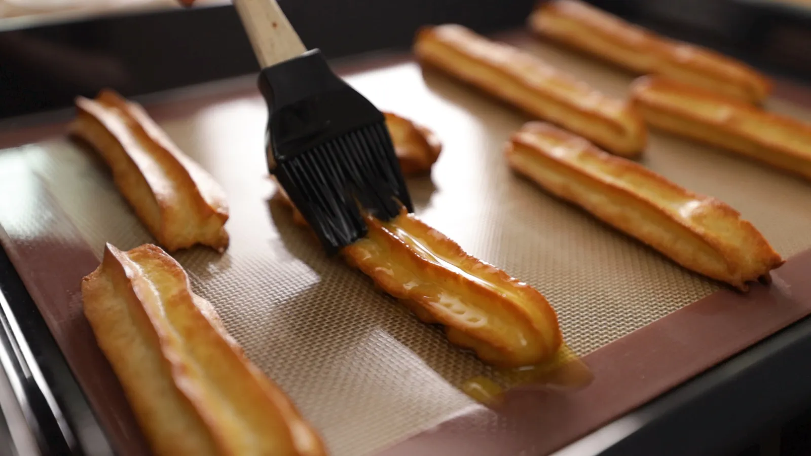 A hand uses a black pastry brush to glaze long, golden-brown choux pastry eclairs on a baking tray lined with a silicone mat.