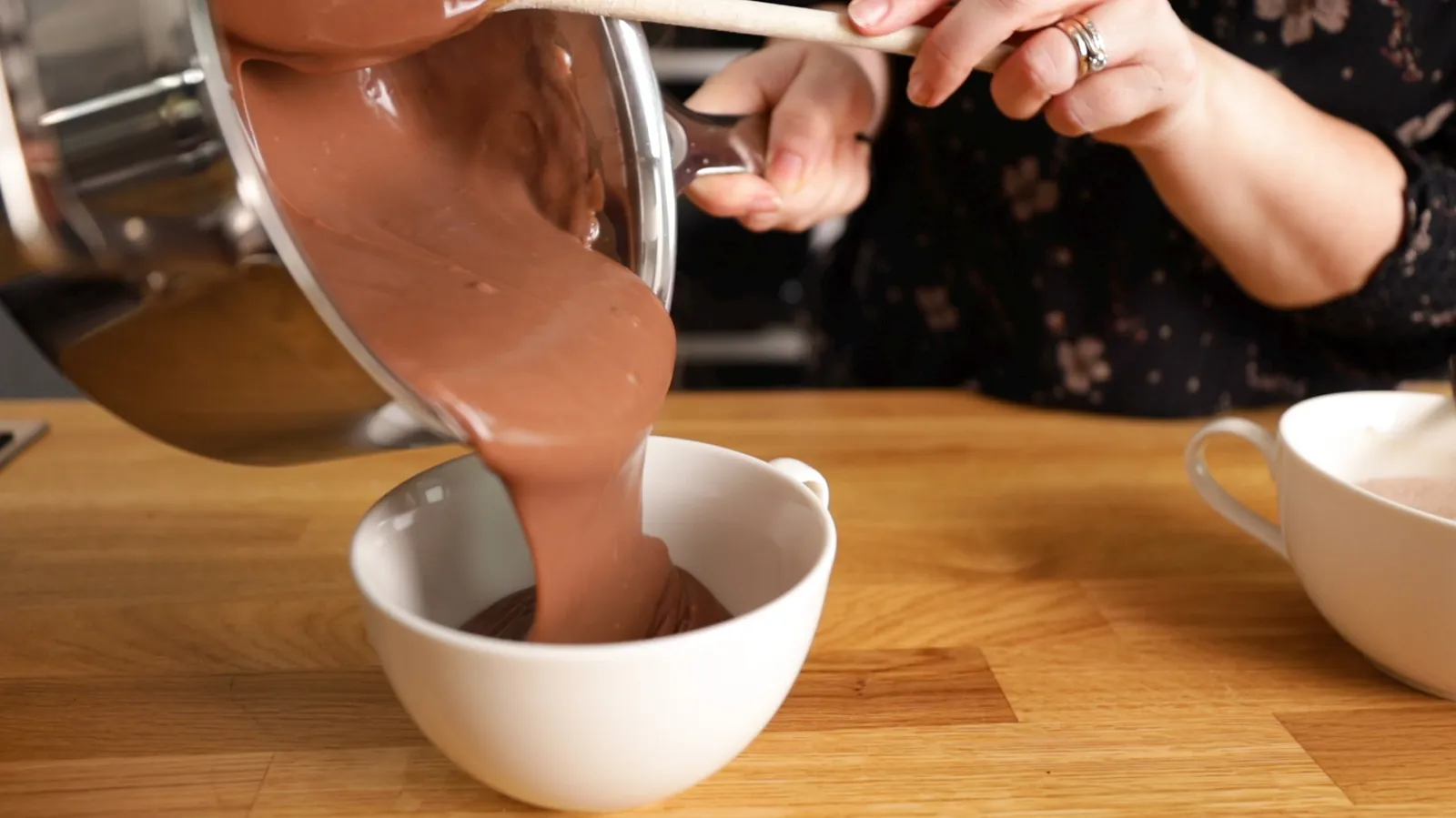 Hands pour thick, light brown chocolate sauce from a stainless steel saucepan into a white ceramic mug.