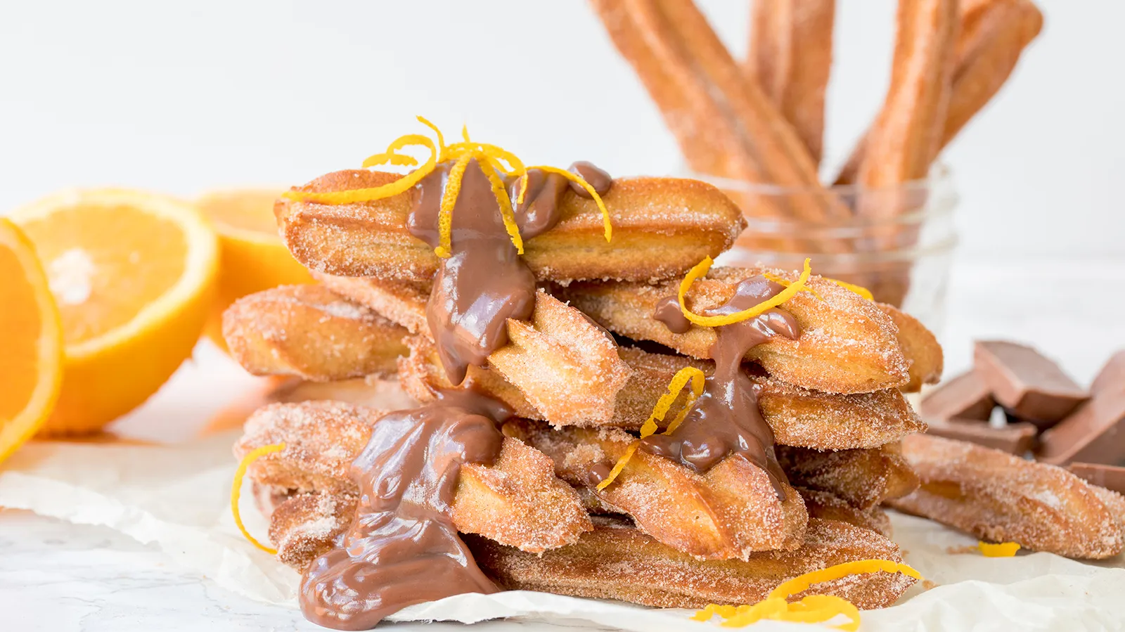 A stack of golden-brown churros, dusted with sugar and cinnamon, drizzled with chocolate sauce and garnished with orange zest, next to sliced oranges and chocolate pieces.