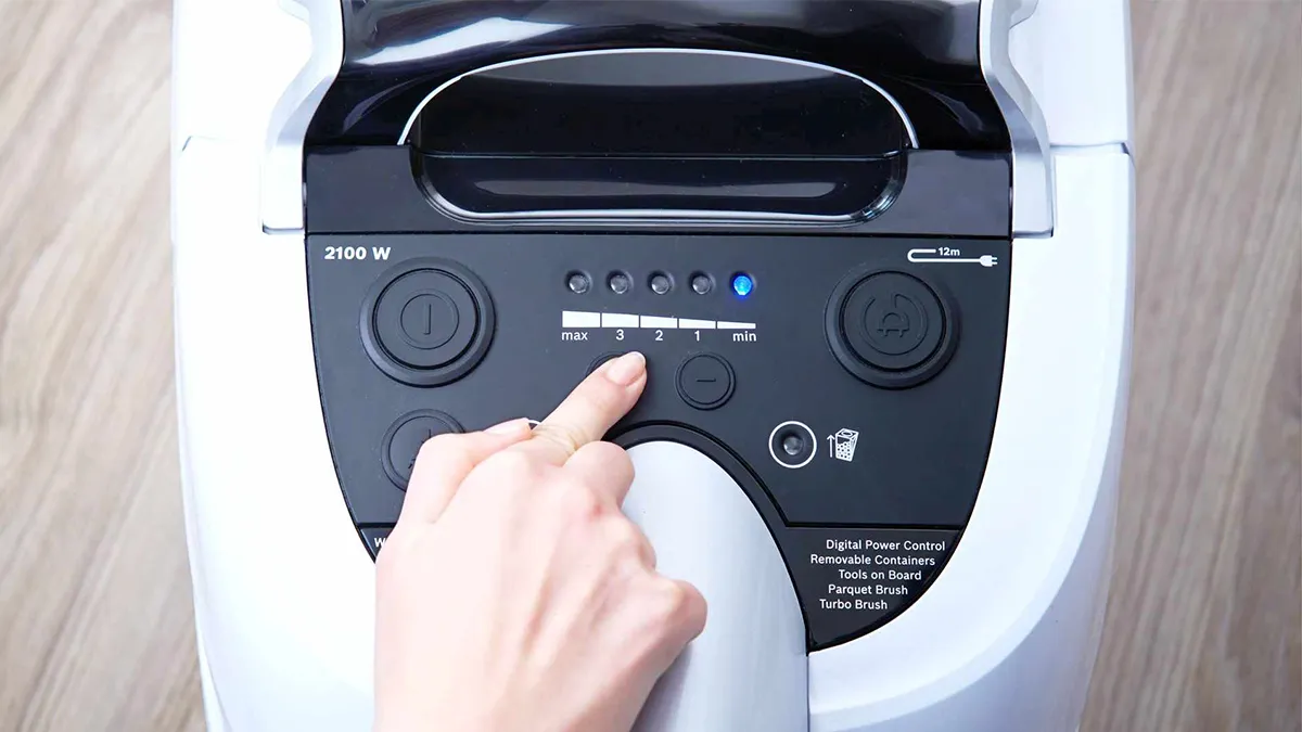 Ręka wskazuje panel sterowania odkurzacza Bosch z mocą 2100 W, zawierający przyciski mocy i zwijania kabla
