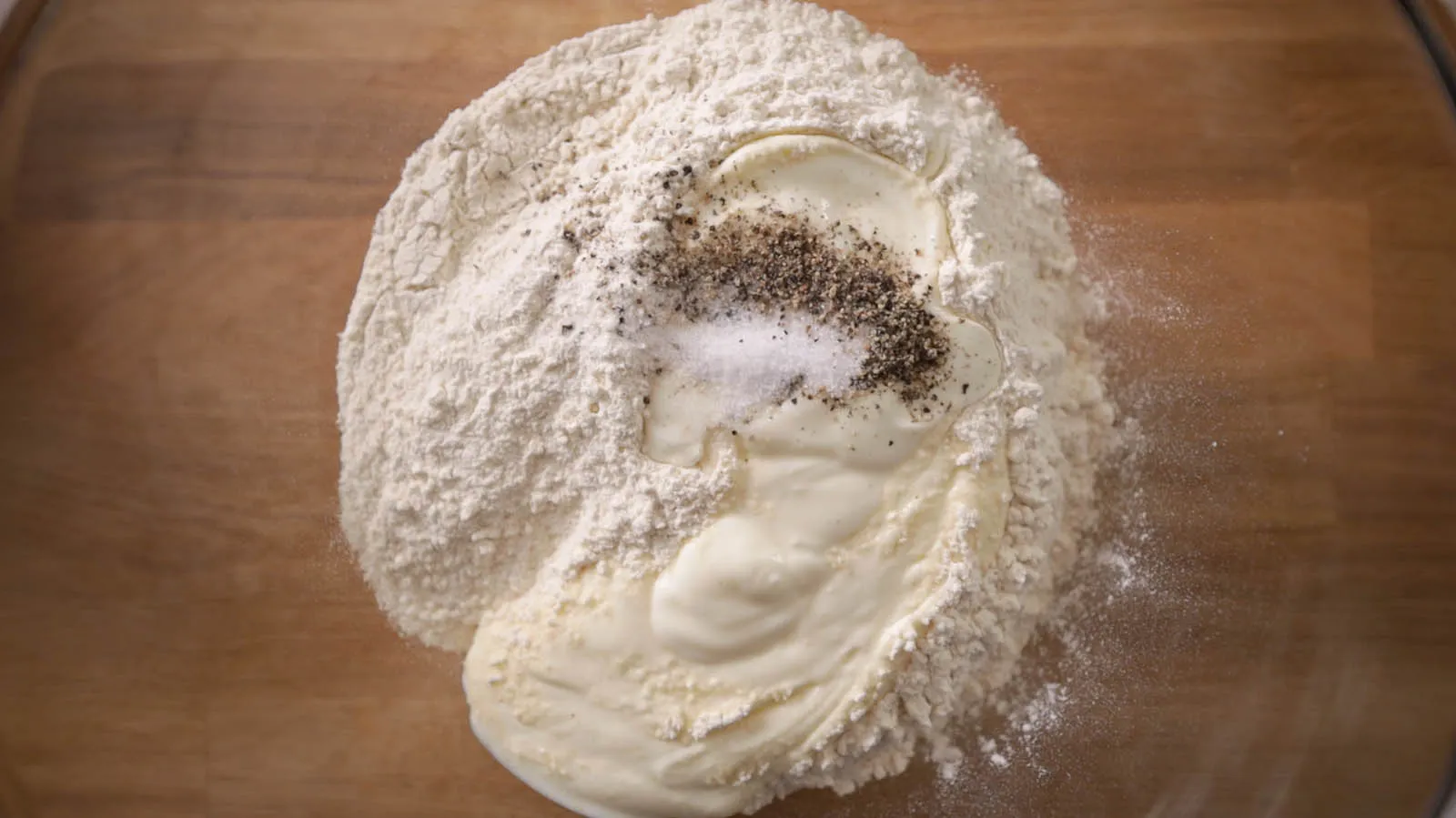 An overhead view of a clear glass bowl containing white flour, a dollop of white cream, salt, and black pepper, ready for mixing.