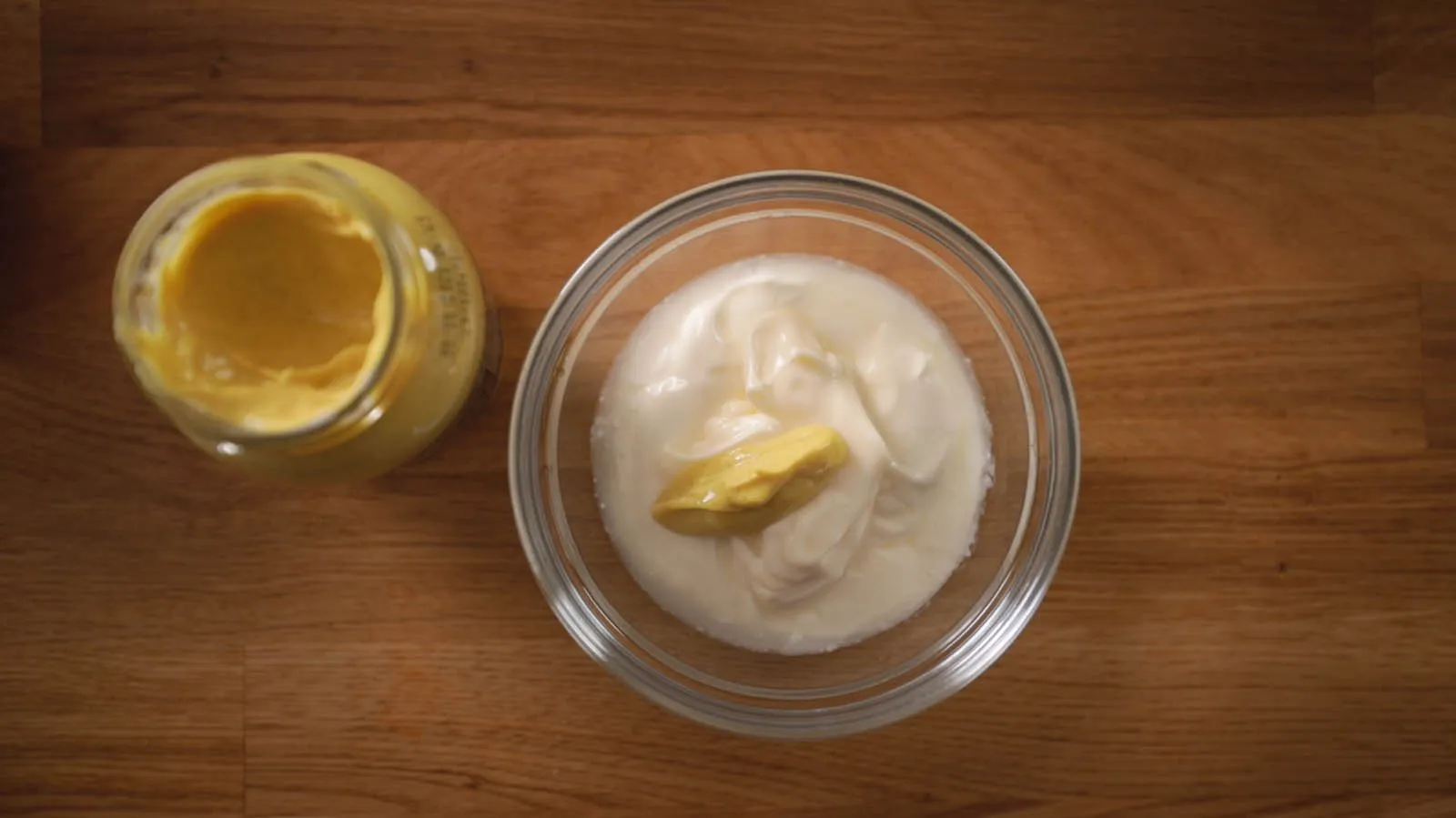 An overhead view of a small glass bowl containing white cream and a dollop of yellow mustard, next to an open jar of mustard, on a wooden countertop.
