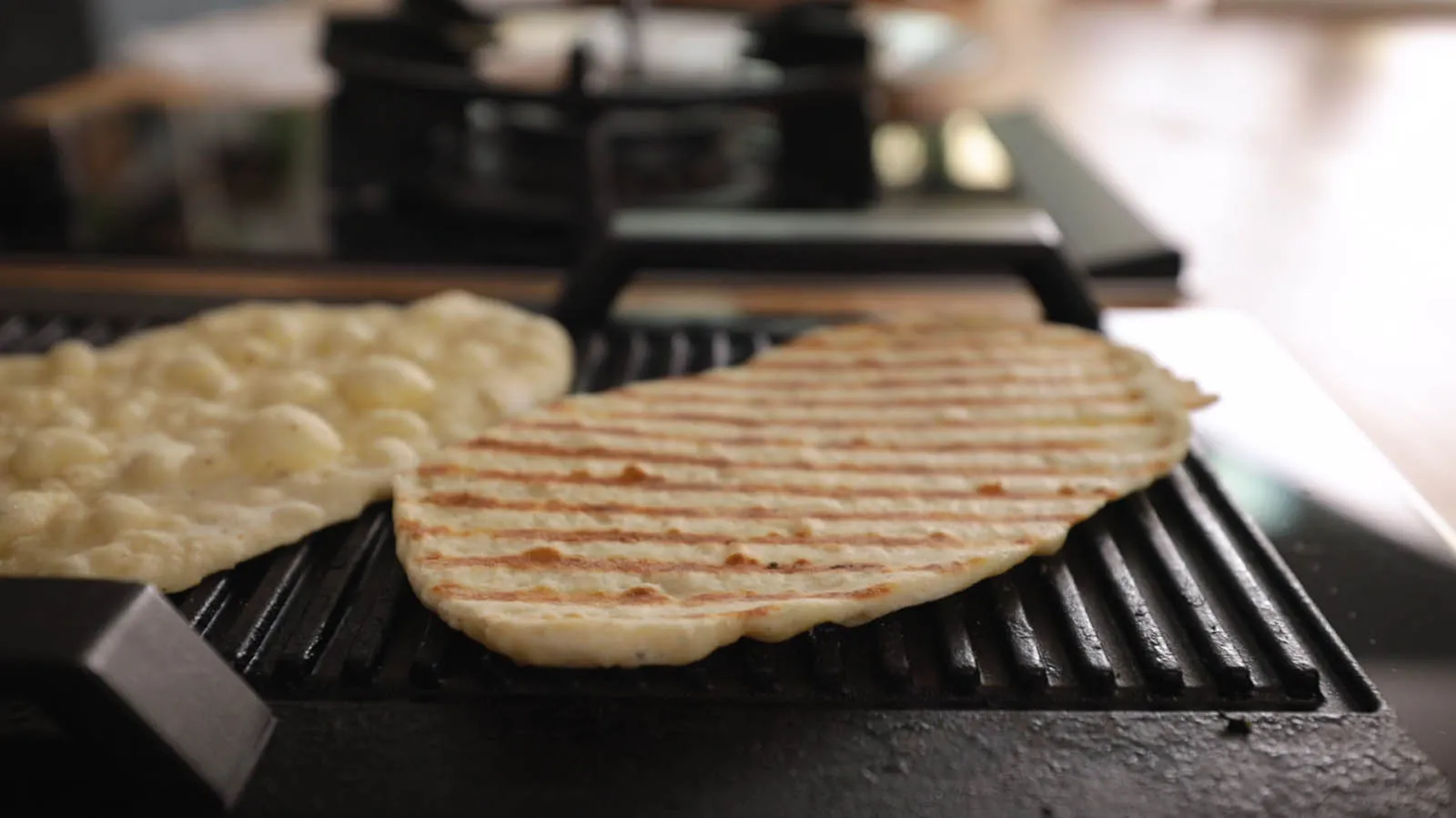 Two pieces of flatbread are cooking on a hot grill pan, one showing prominent grill marks.