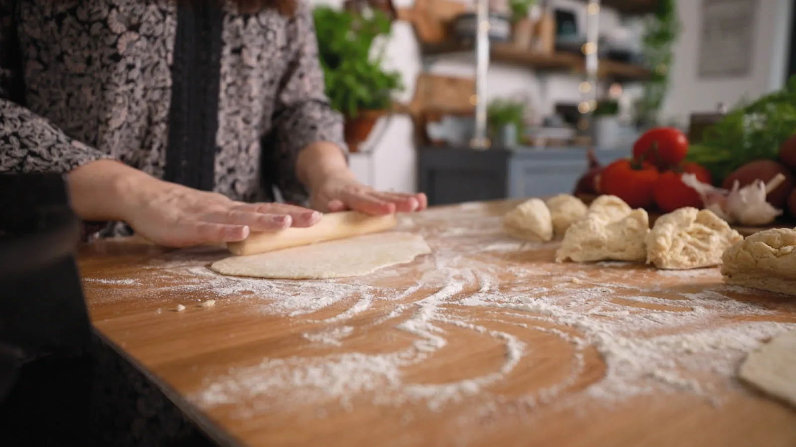 Hands use a wooden rolling pin to flatten a piece of dough on a floured wooden countertop, with other dough portions nearby.