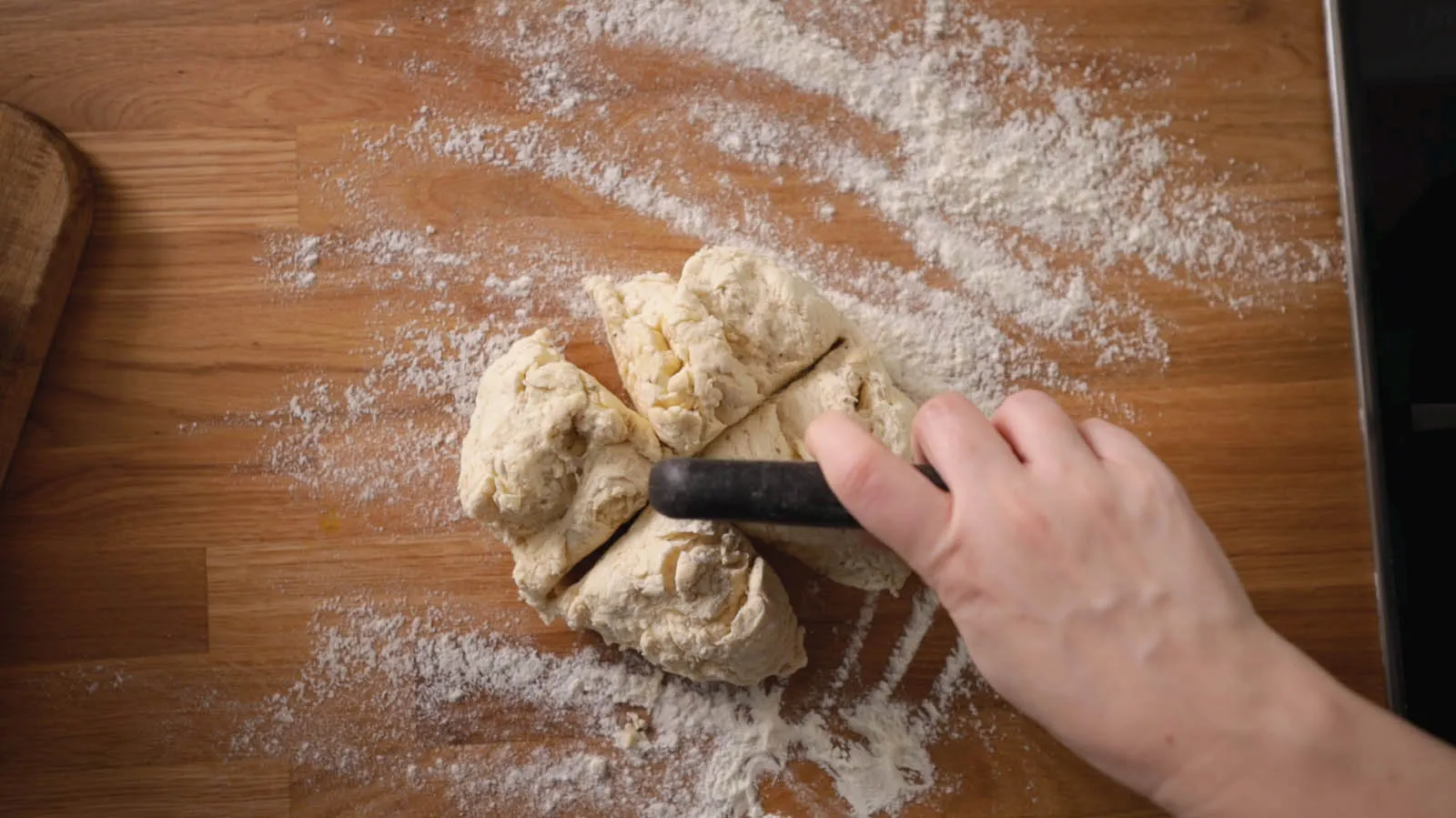 An overhead view of a hand using a dough cutter to divide a ball of dough into several portions on a floured wooden countertop.
