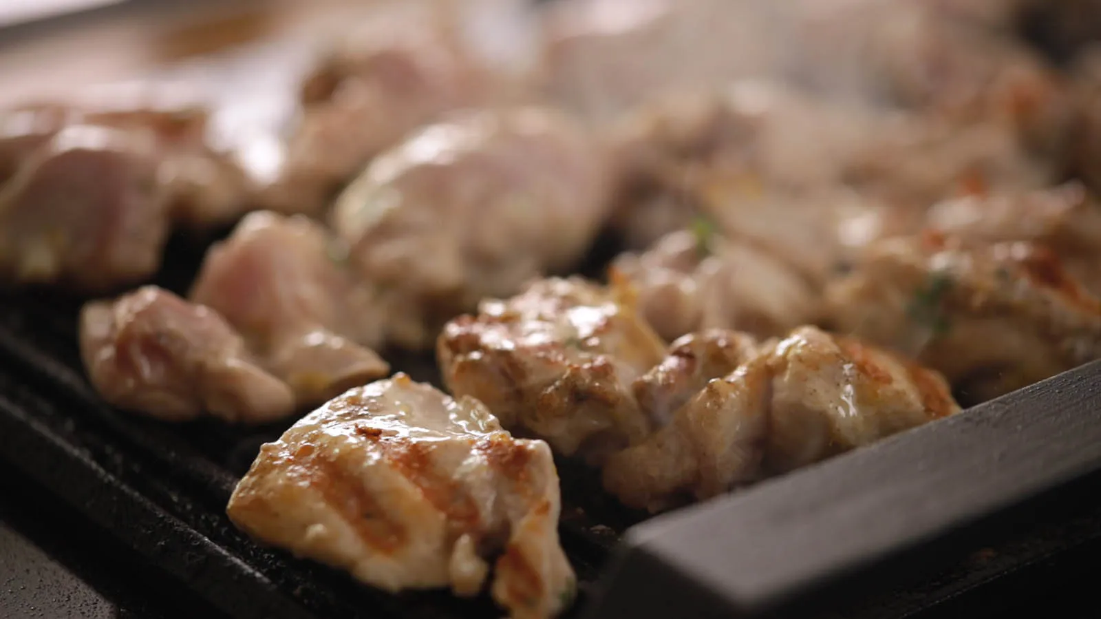 A close-up of chicken pieces sizzling and smoking on a hot grill pan, showing grill marks.