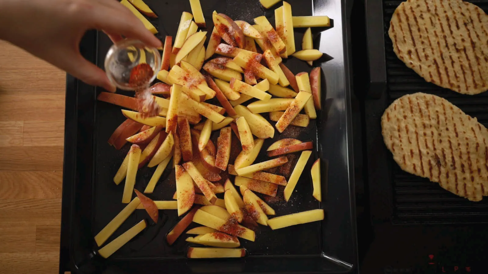 A hand sprinkles red seasoning from a small glass shaker over a baking tray filled with raw potato fries, next to grilled flatbreads.