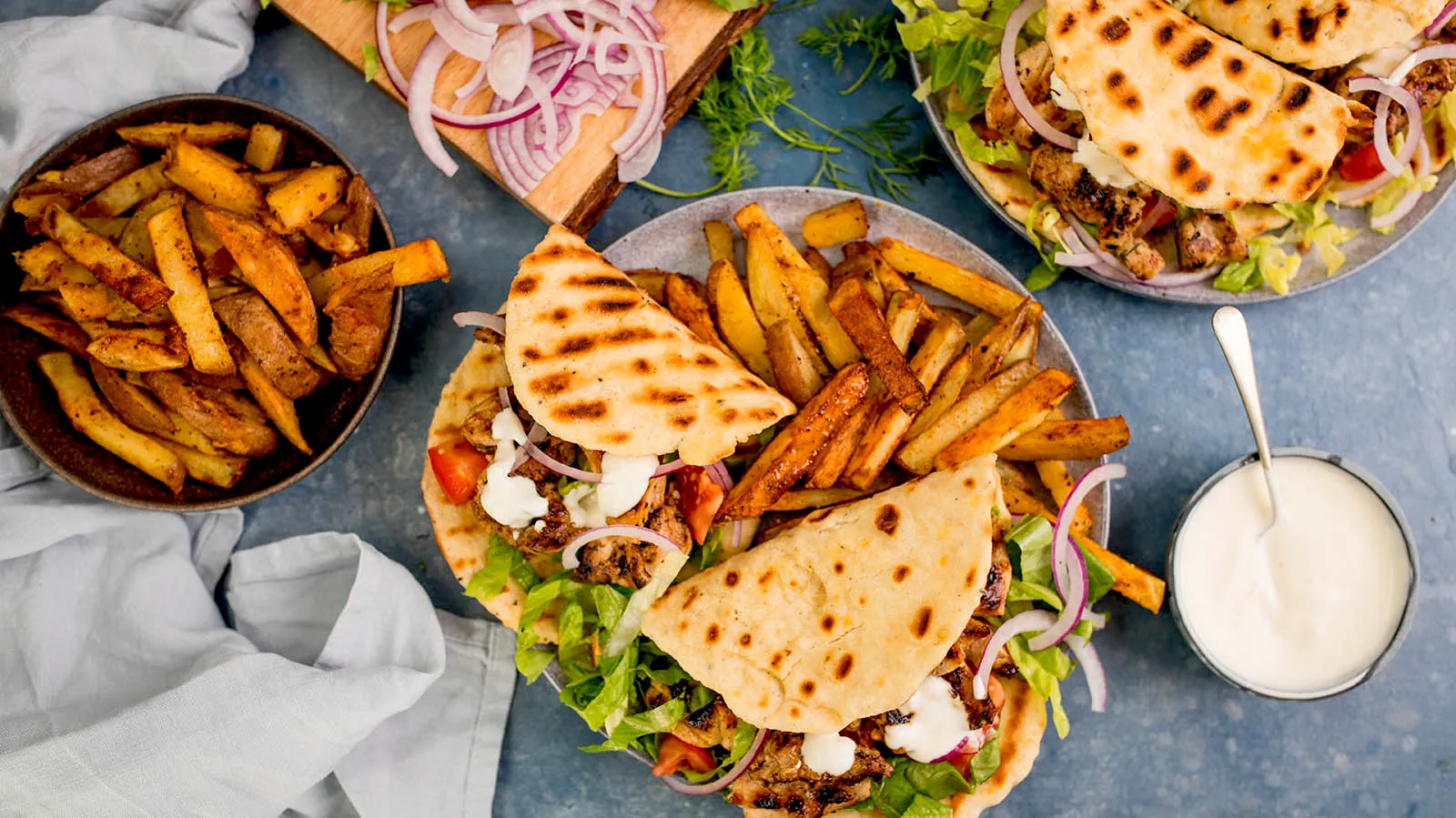 An overhead view of two plates of chicken gyros with grilled flatbread, lettuce, red onion, tomatoes, and a white sauce, served with a side of seasoned fries and a bowl of white sauce.