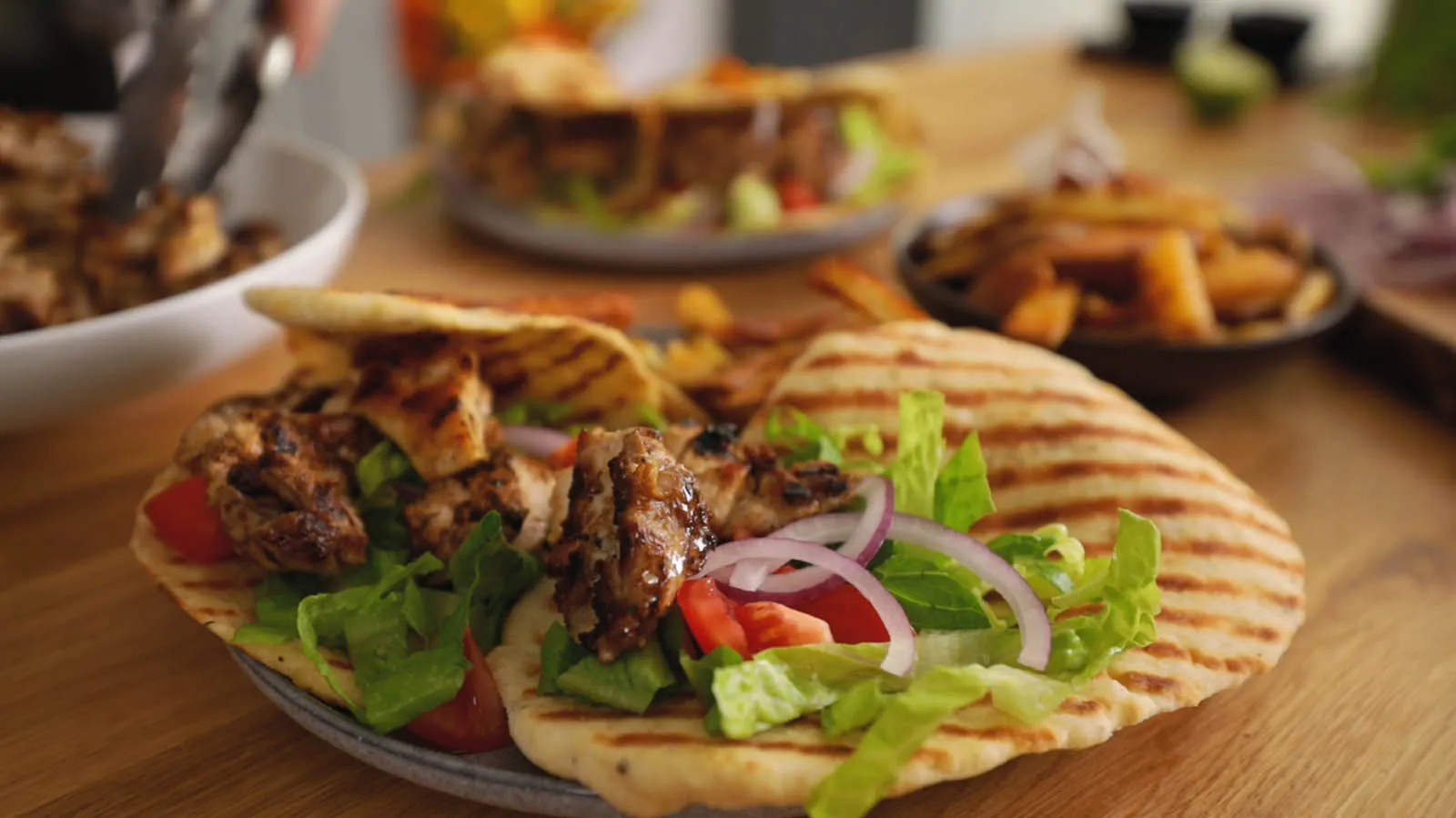 A hand uses tongs to add grilled chicken pieces to a plate with grilled flatbread, lettuce, red onion, and tomatoes, assembling a gyro.