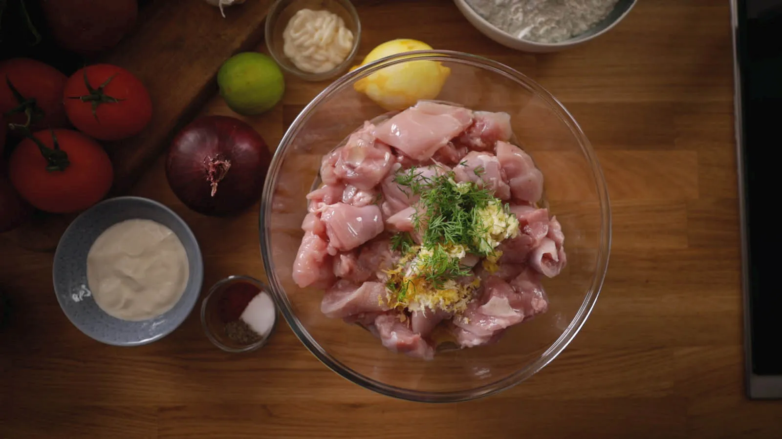 An overhead view of a clear glass bowl filled with diced raw chicken, minced garlic, lemon zest, and fresh dill, surrounded by various ingredients like tomatoes, onions, and spices on a wooden countertop.