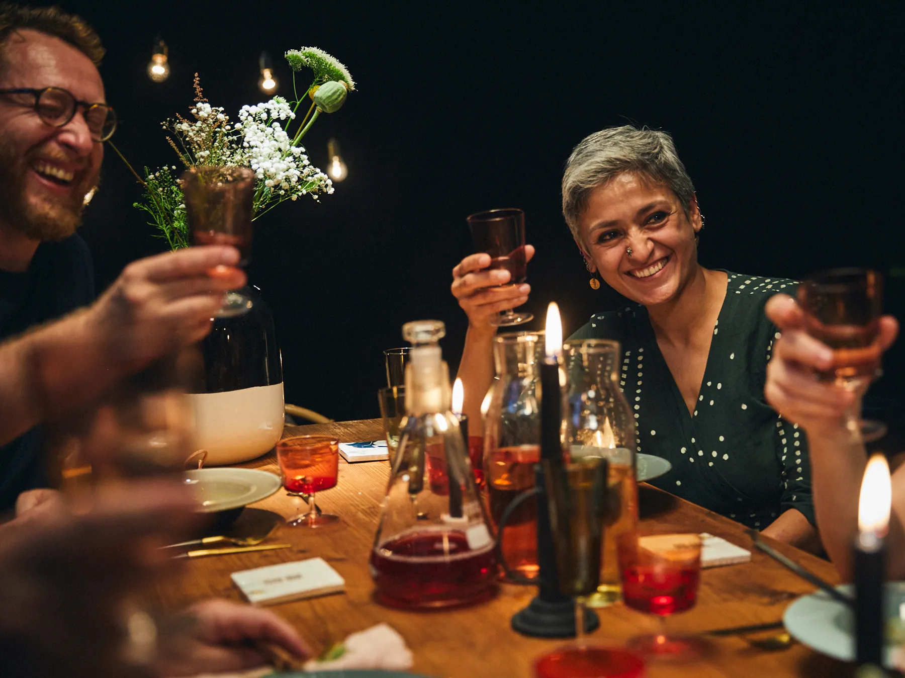 Cena festa con amici, bevande, fiori e risate in un ambiente accogliente e illuminato.