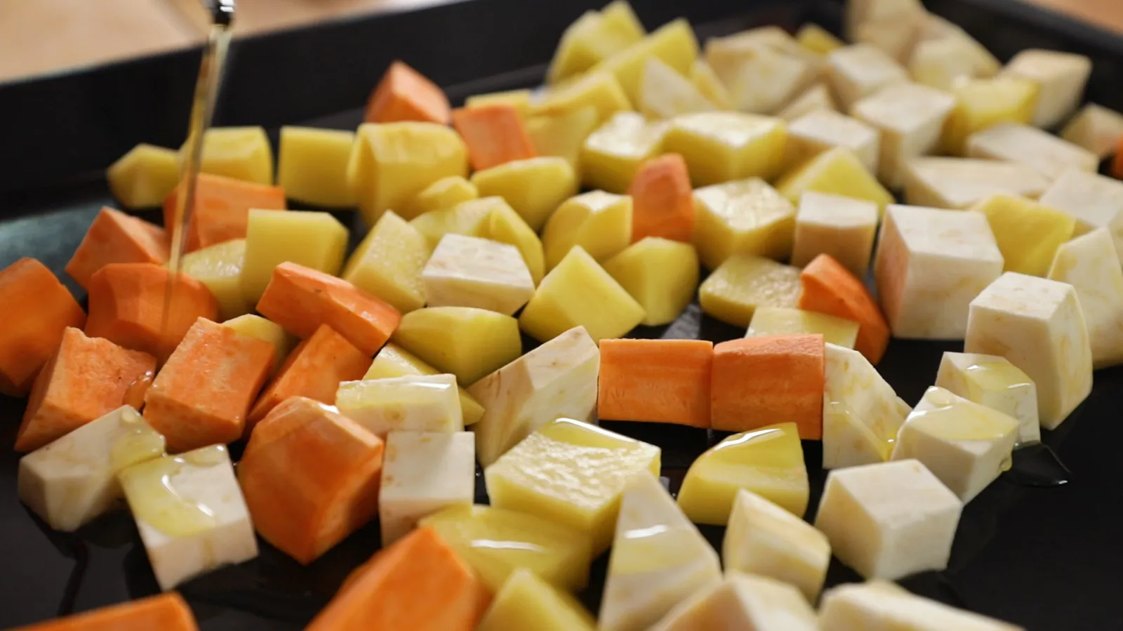 A close-up of a baking tray filled with diced root vegetables (potatoes, sweet potatoes, parsnips) being drizzled with olive oil.