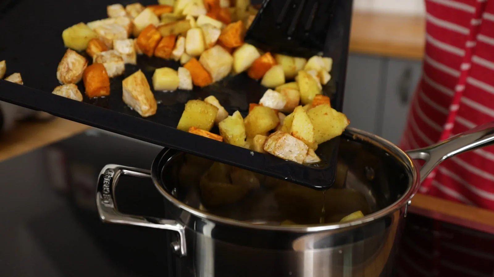 A black spatula slides roasted root vegetables and apples from a baking tray into a stainless steel pot on an induction cooktop.