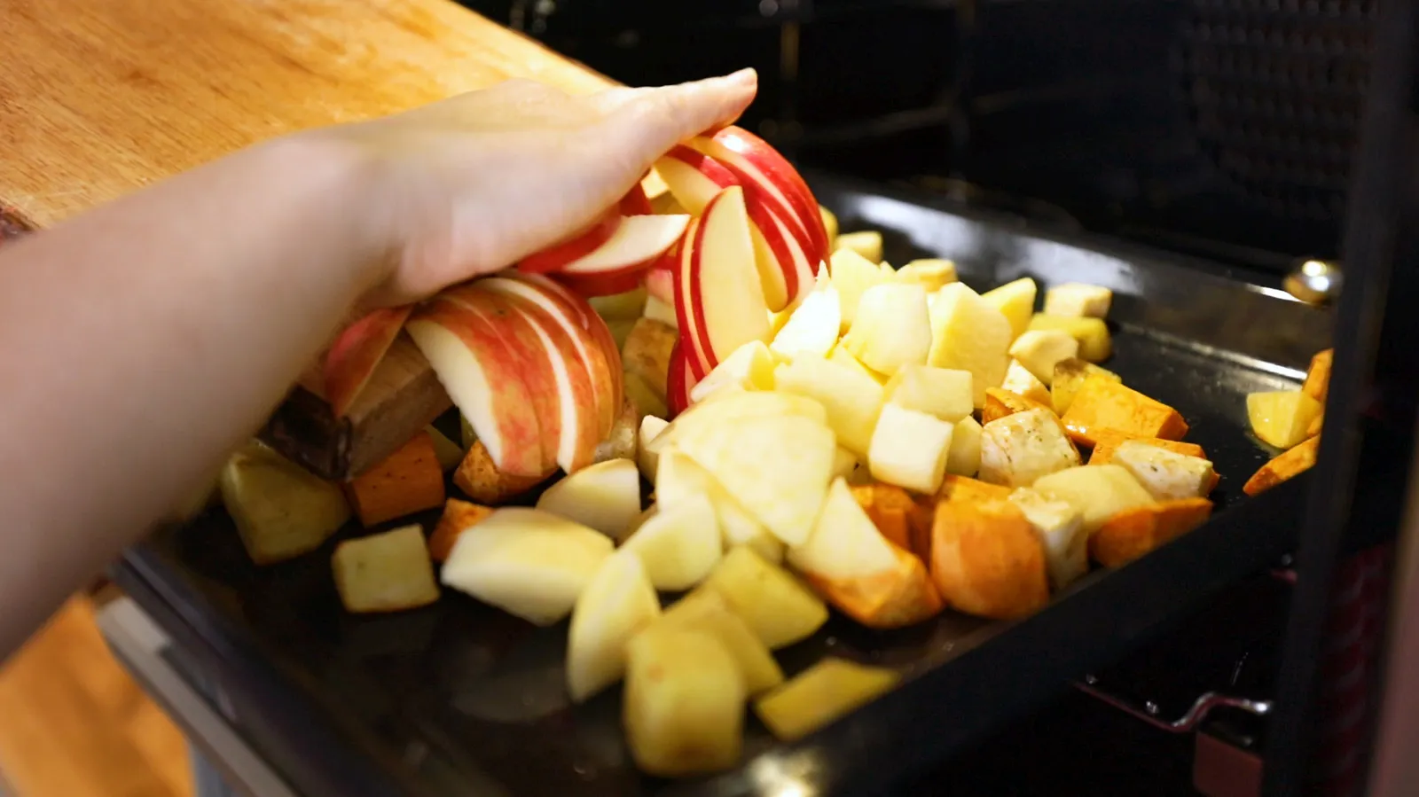 A hand adds sliced red apples to a baking tray filled with diced root vegetables, ready for roasting