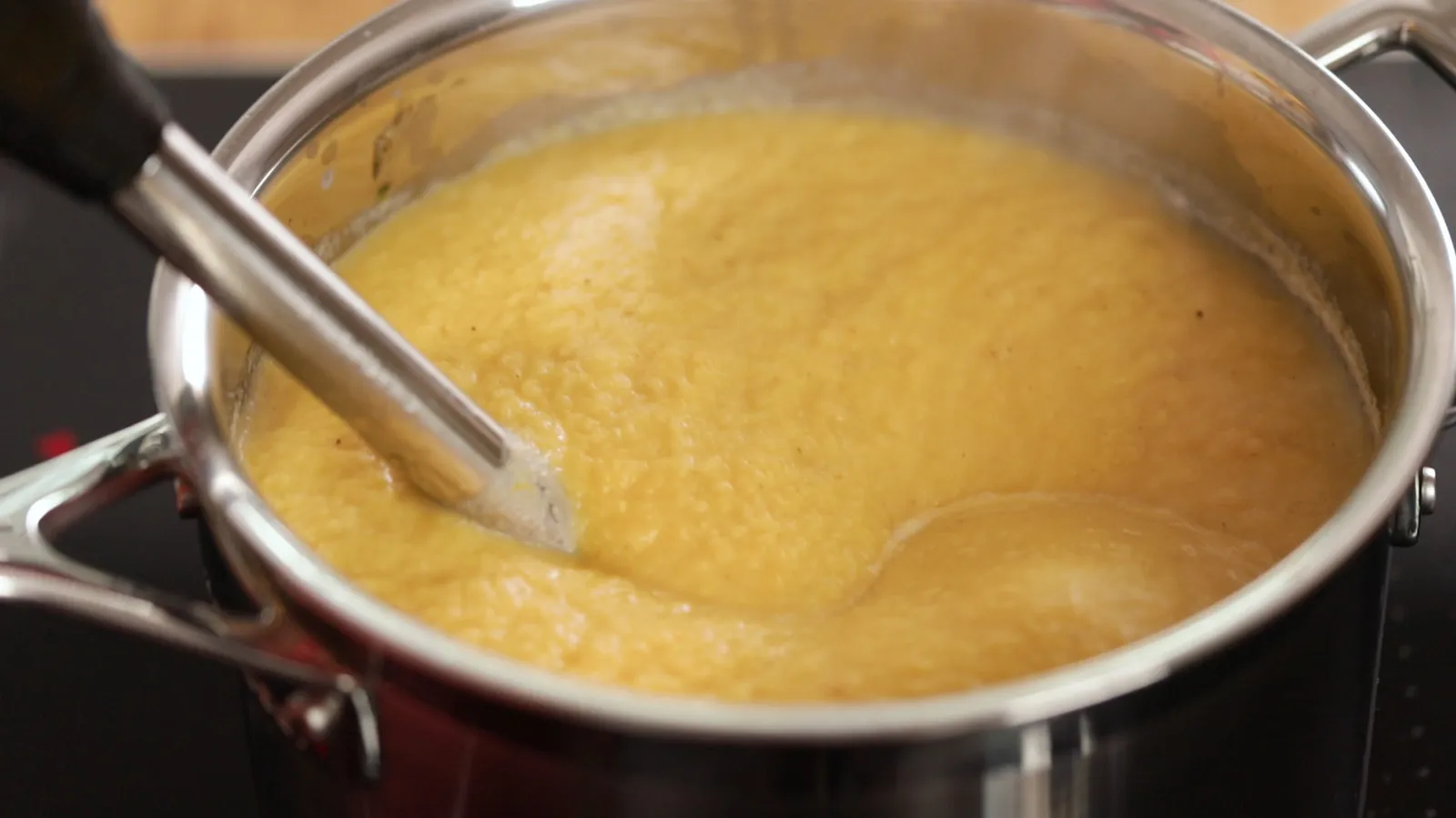 A close-up of a stainless steel pot on an induction cooktop, filled with simmering orange soup, being blended with an immersion blender.