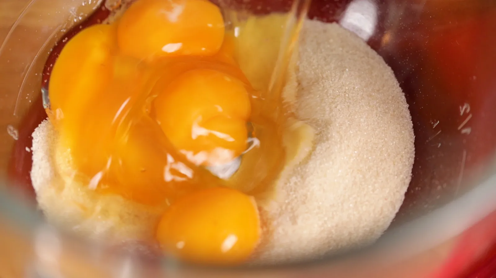 A close-up of a clear glass mixing bowl containing several egg yolks and white granulated sugar.