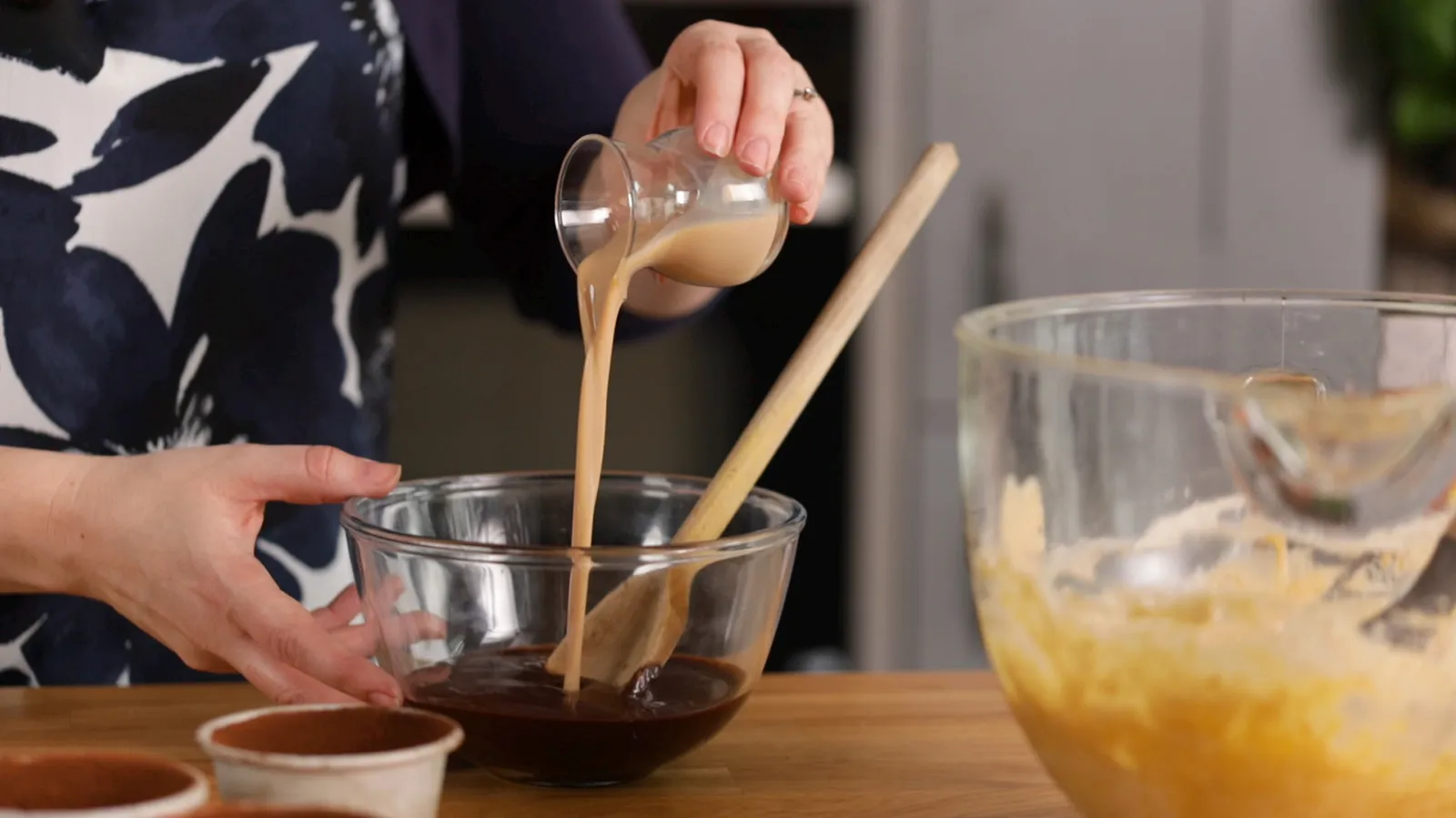 A hand pours a stream of light brown liquid (likely coffee or liqueur) from a small glass into a clear glass bowl containing melted chocolate, next to a wooden spoon.