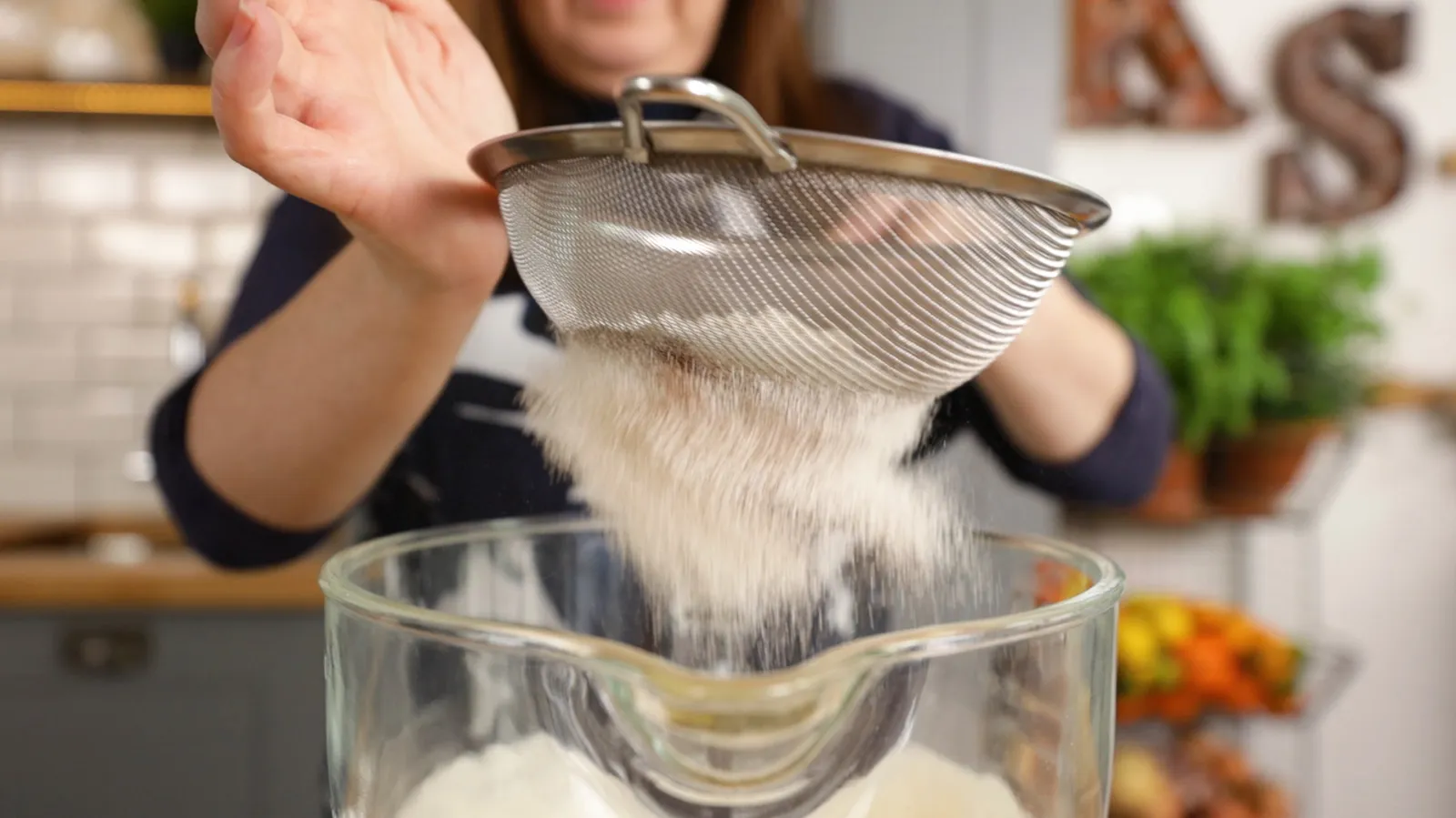 A hand sifts white flour from a metal sieve into a large clear glass mixing bowl.