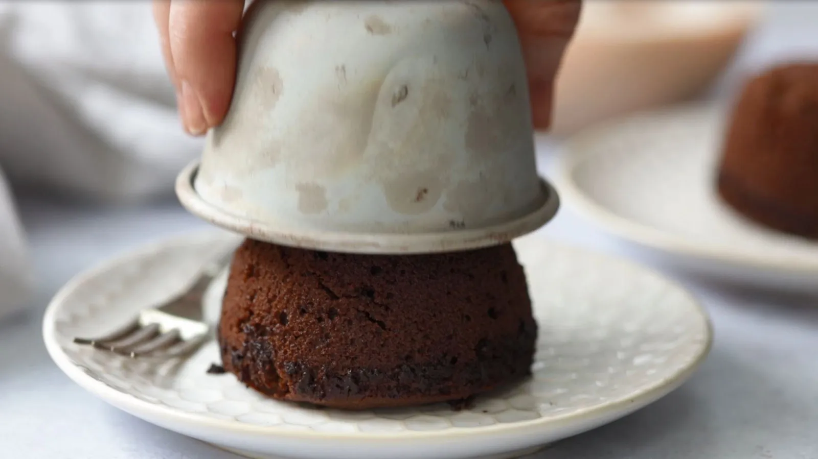 A hand lifts a small, light grey ceramic bowl from an inverted chocolate lava cake on a white plate, revealing the perfectly formed cake.