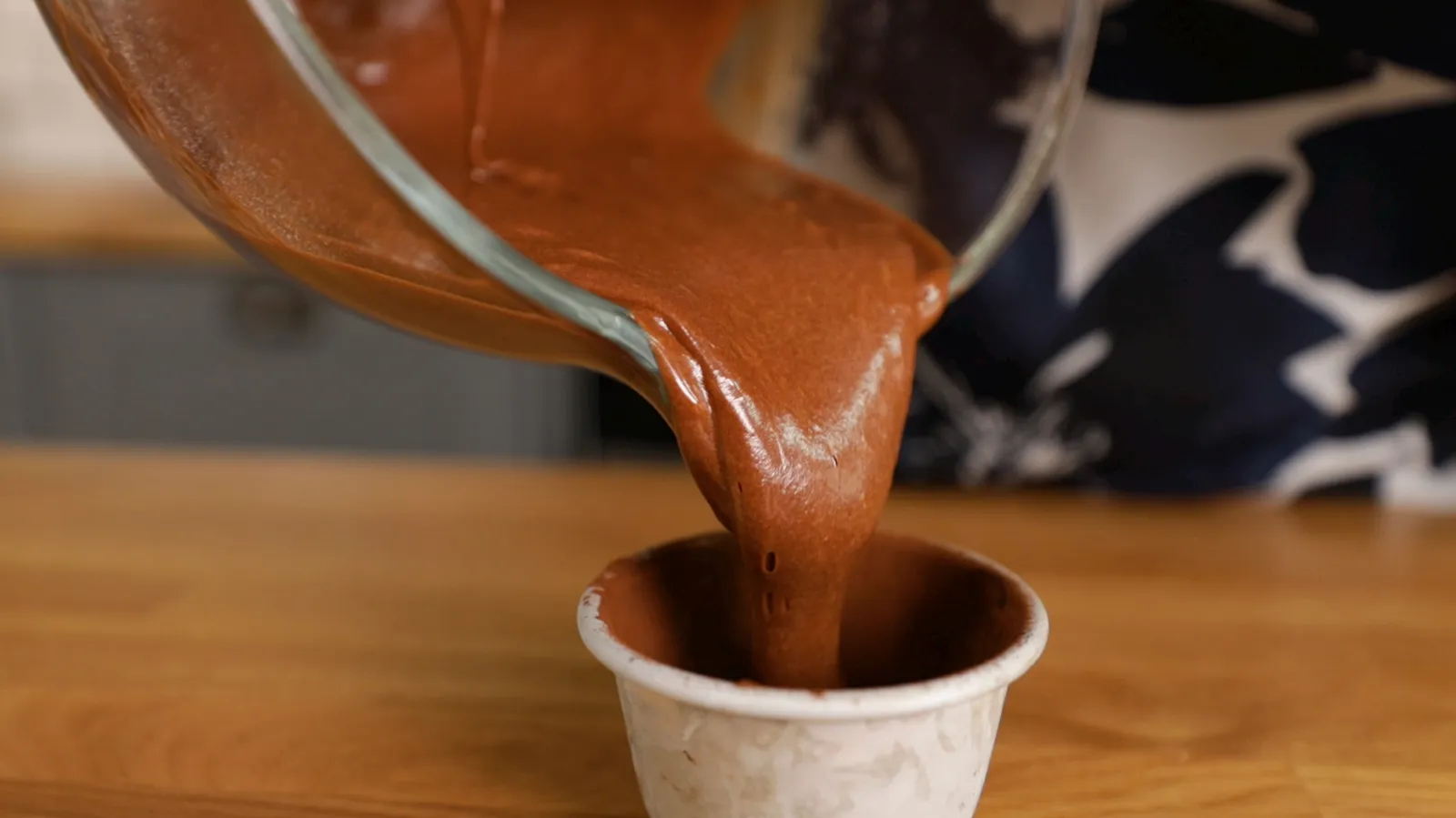 Thick, dark chocolate batter is poured from a clear glass bowl into a small white paper baking cup.