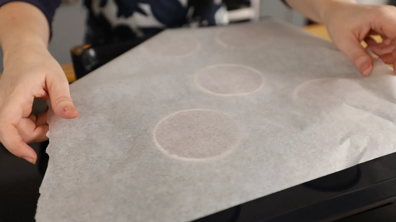 Hands place a sheet of parchment paper with circular outlines onto a baking tray, preparing for baking.