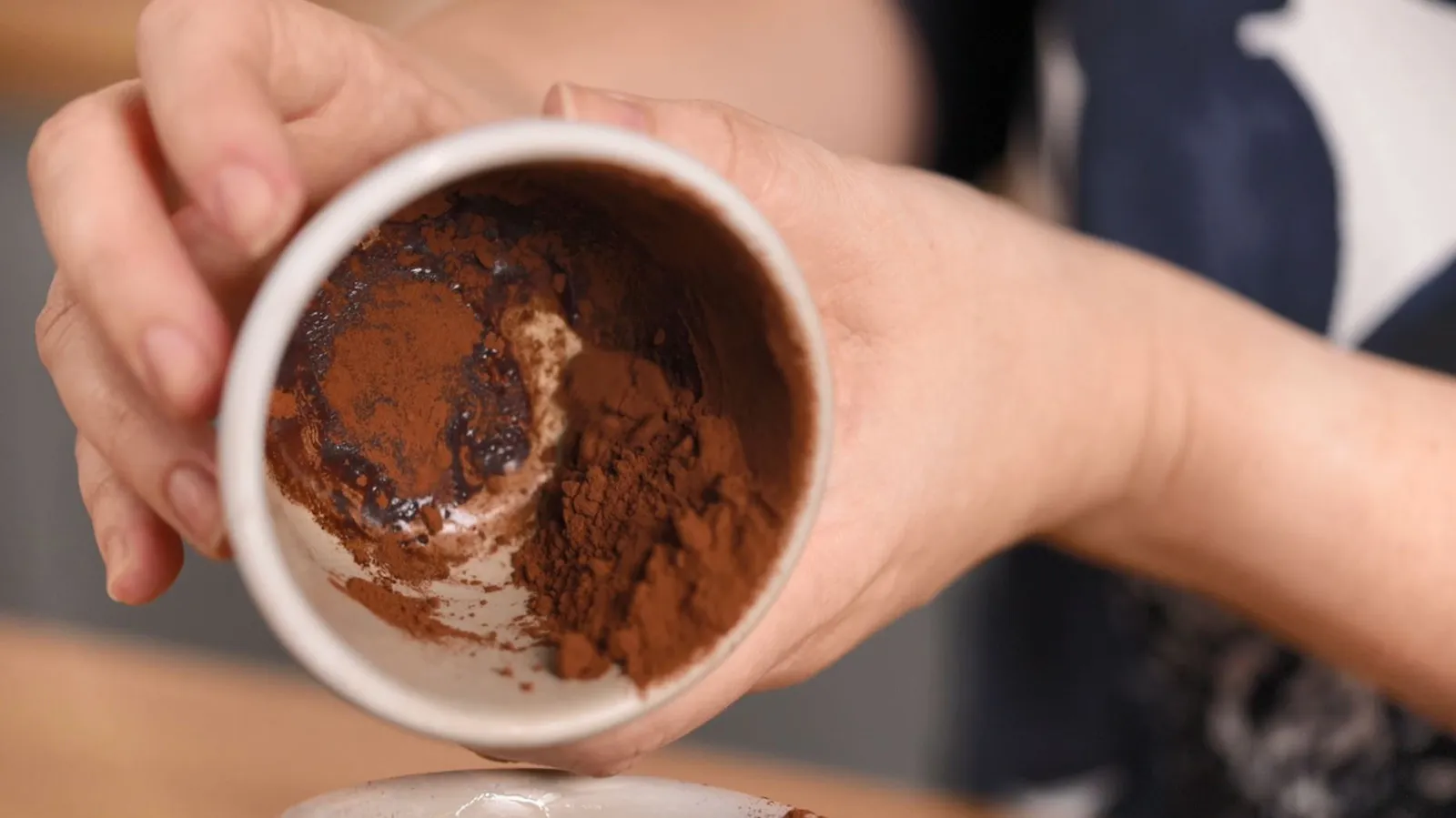 Hands hold a small white cup, tilted to show a mixture of dark cocoa powder and a dark liquid (possibly coffee or melted chocolate) inside.