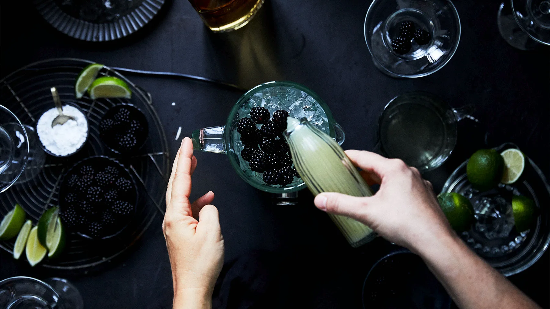An overhead shot of hands preparing a cocktail, pouring a light green liquid into a pitcher with ice and blackberries, surrounded by limes, sugar, and glasses on a dark surface.