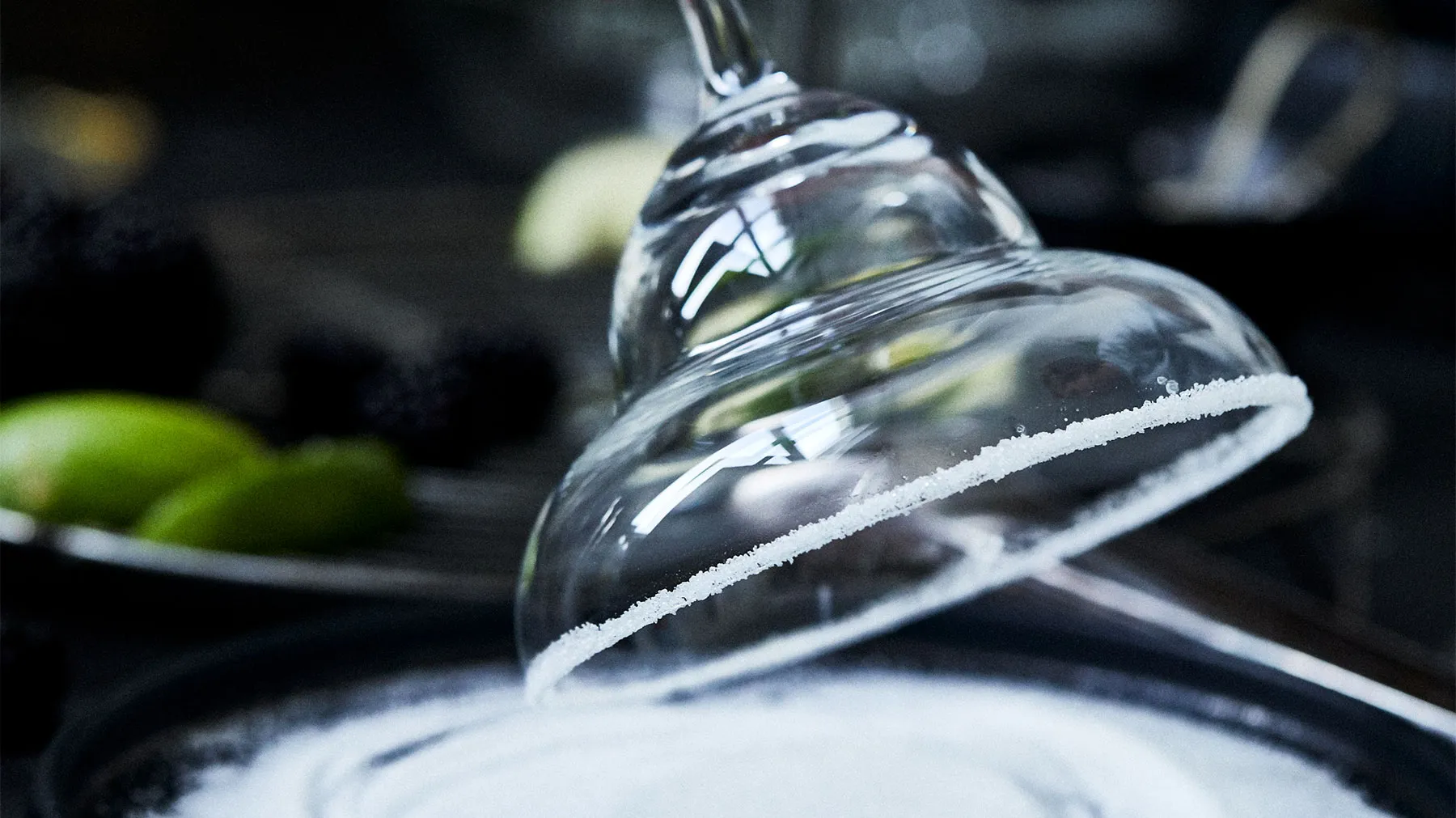 A margarita glass with a salted rim is inverted over a pile of white salt, with limes and blackberries in the background.