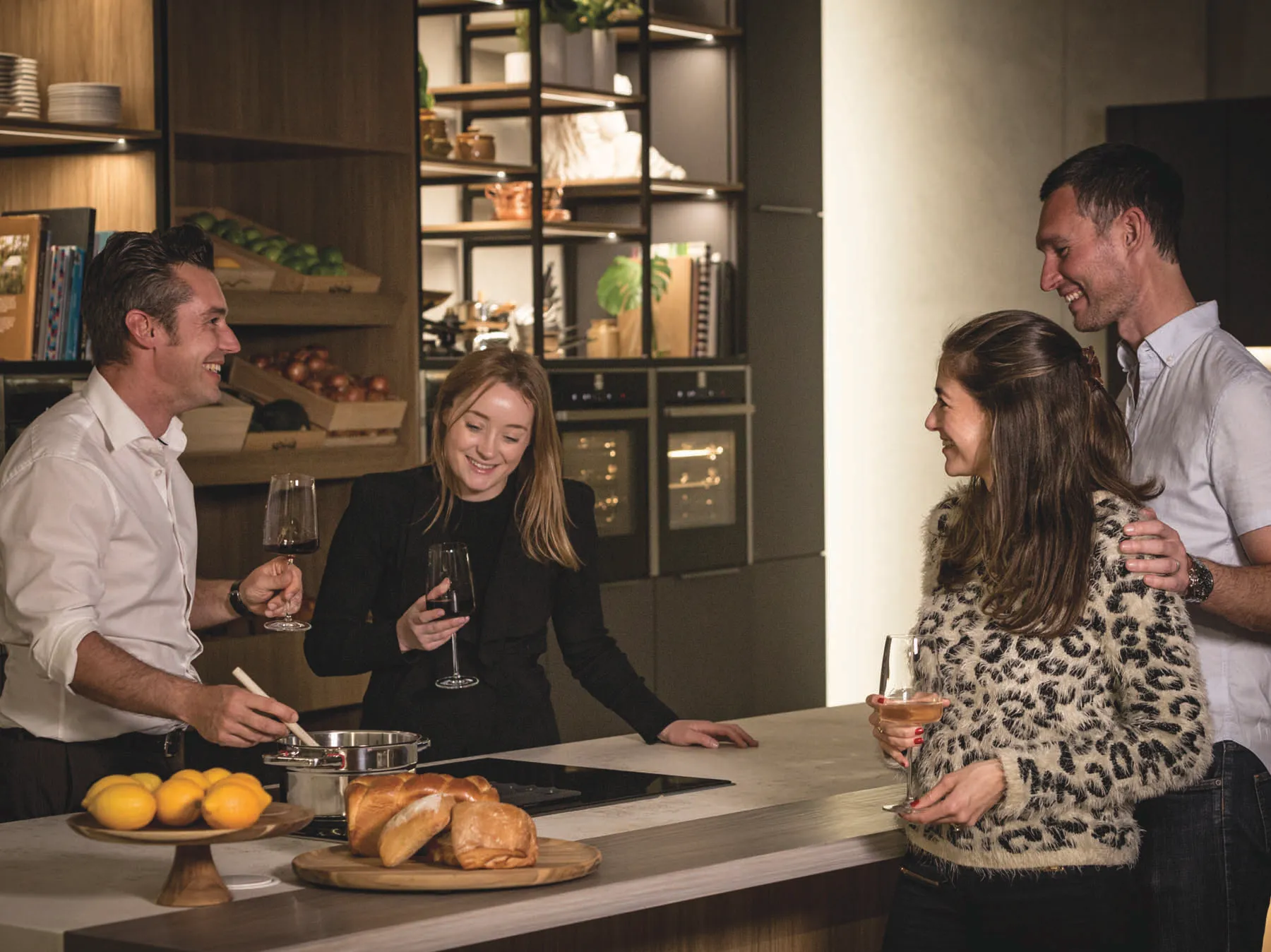 Four adults are socializing in a modern, well-appointed kitchen. A man in a white shirt stirs a pot on a sleek induction cooktop, holding a glass of red wine and smiling. Next to him, a woman in a black blazer also smiles while holding a glass of red wine. On the right, another woman in a fluffy leopard print sweater holds a cocktail, while a man with his arm around her smiles warmly. The light-colored countertop features a wooden stand with lemons and a cutting board with fresh bread rolls. In the background, dark wooden shelving units display books, plants, and decorative items, with modern built-in ovens visible. The group is laughing and engaged in conversation, creating a warm, social atmosphere.