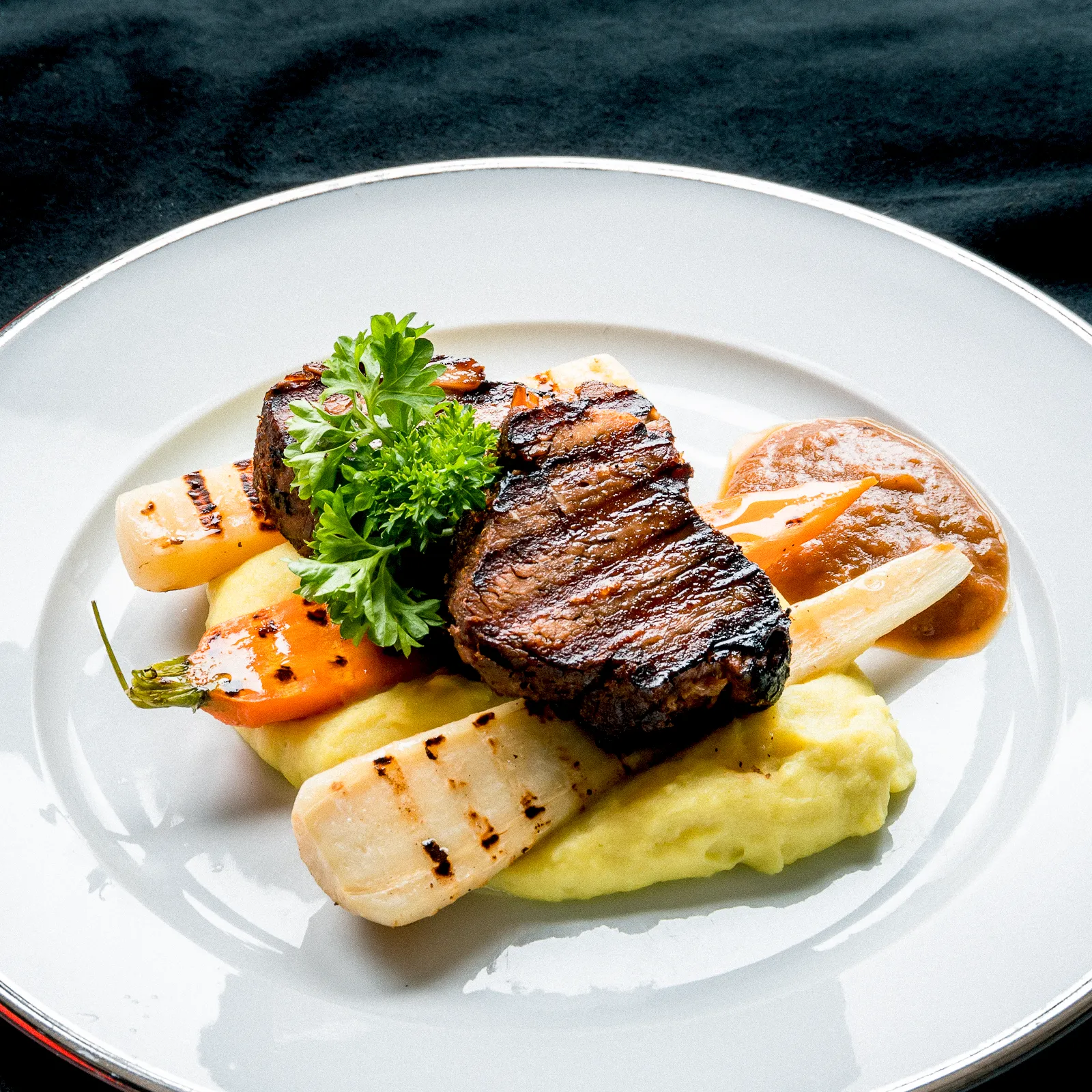 Ψητή μπριζόλα με ψητά λαχανικά και πουρέ πατάτας σε λευκό πιάτο.