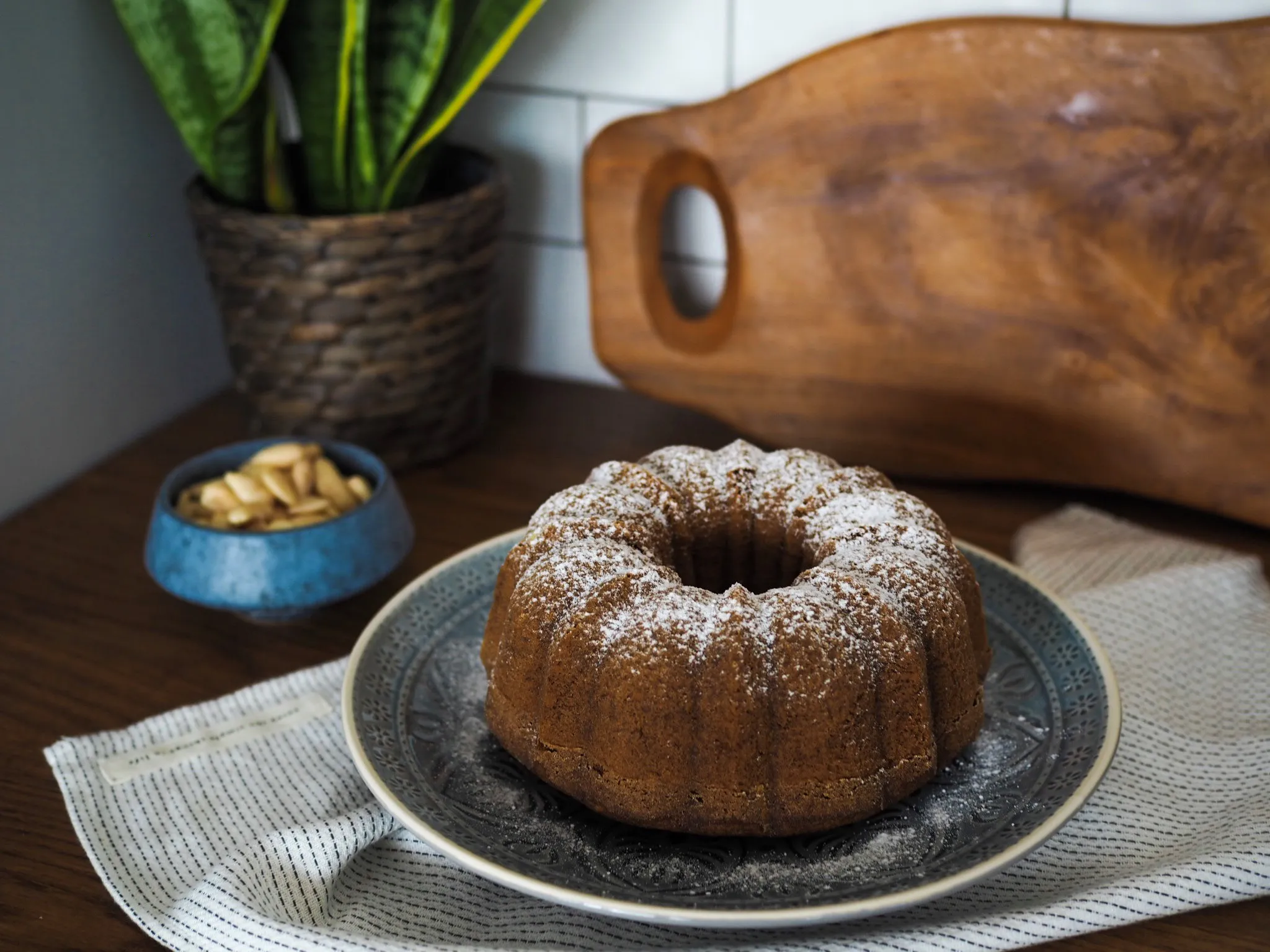 Čerstvo pečený bundt torta oprášená práškovým cukrom, obklopená kuchynským náčiním a zeleňou.