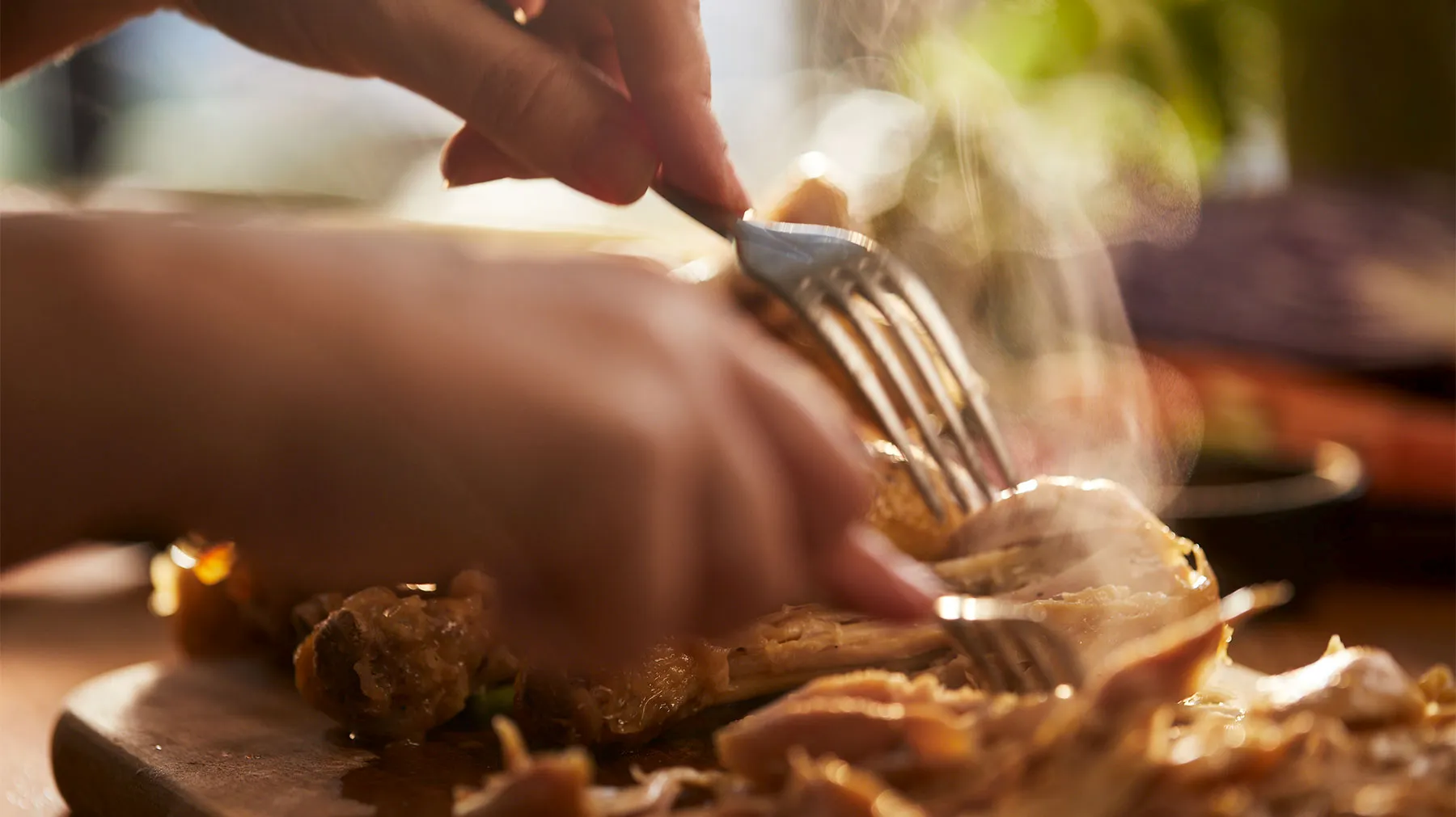 Hands using fork to cut into cooked meat dish on wooden surface, with steam rising from the food.