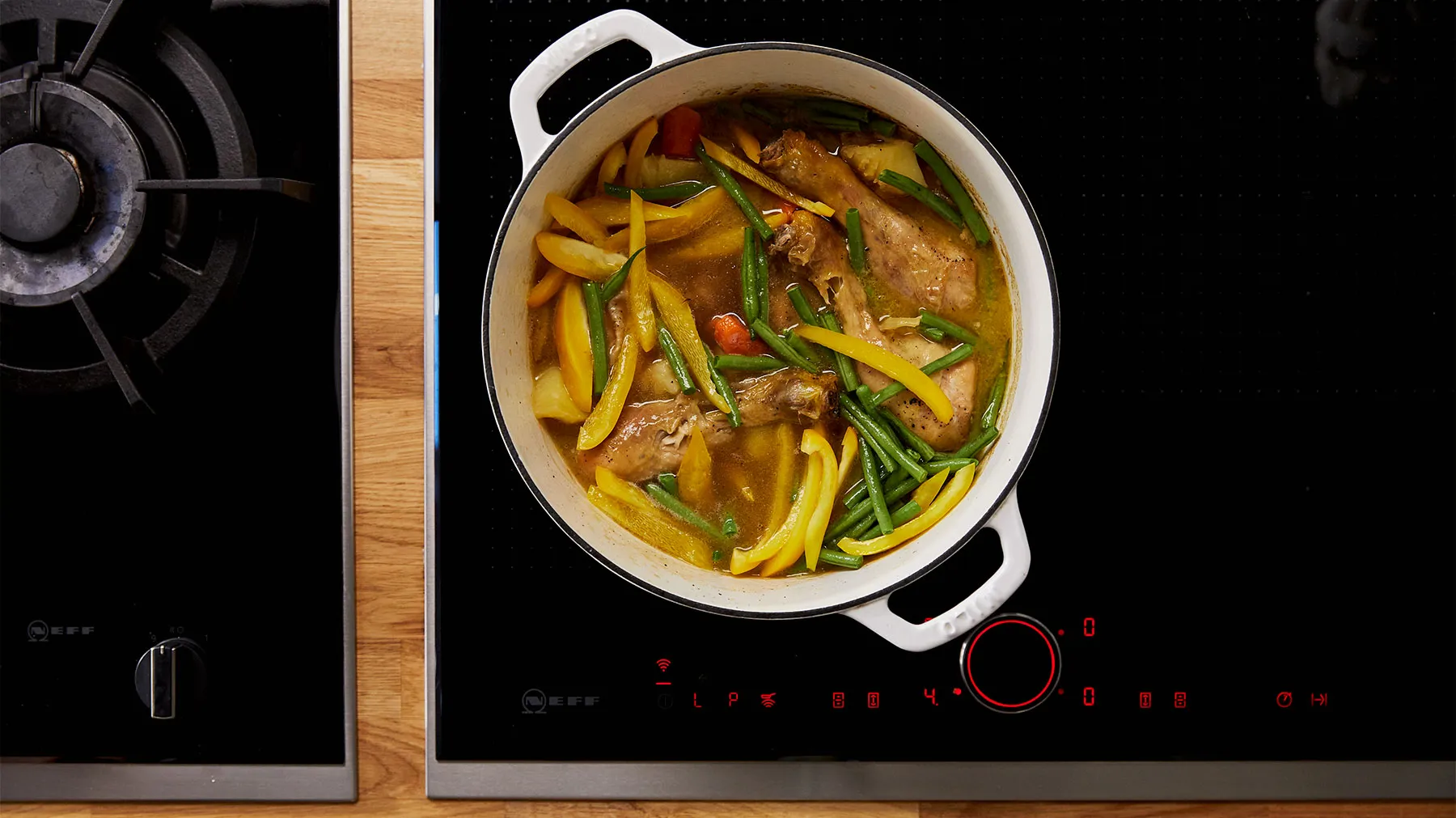 An overhead view of a white enameled cast iron pot filled with chicken, potatoes, green beans, and yellow bell peppers in a broth, simmering on a black induction cooktop.