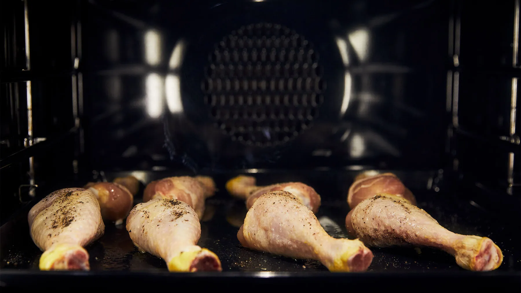 Raw chicken drumsticks, seasoned with salt and pepper, are arranged on a black baking tray inside a dark oven.