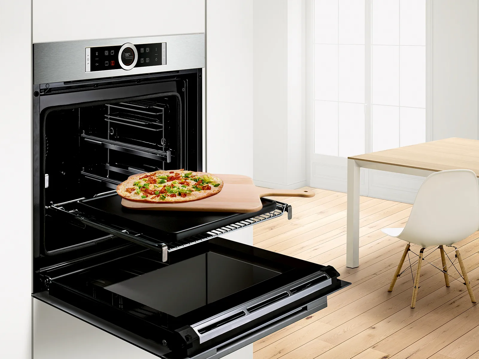 Moderna kuhinja s pećnicom, pizza na plehu, drveni stol i stolica u pozadini.
