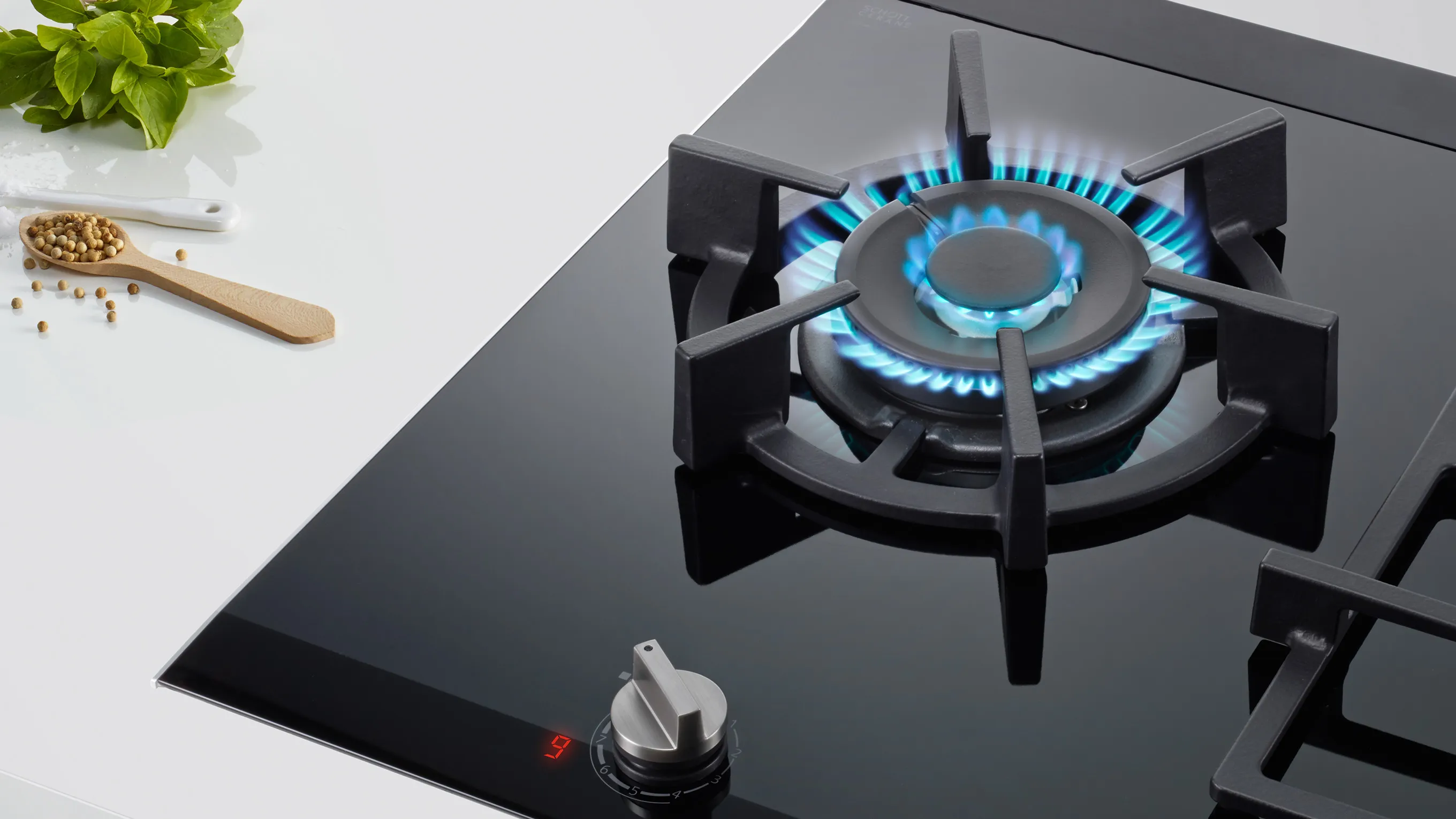 Black gas stove burner with blue flames, cooking utensils, and green plant on white surface.
