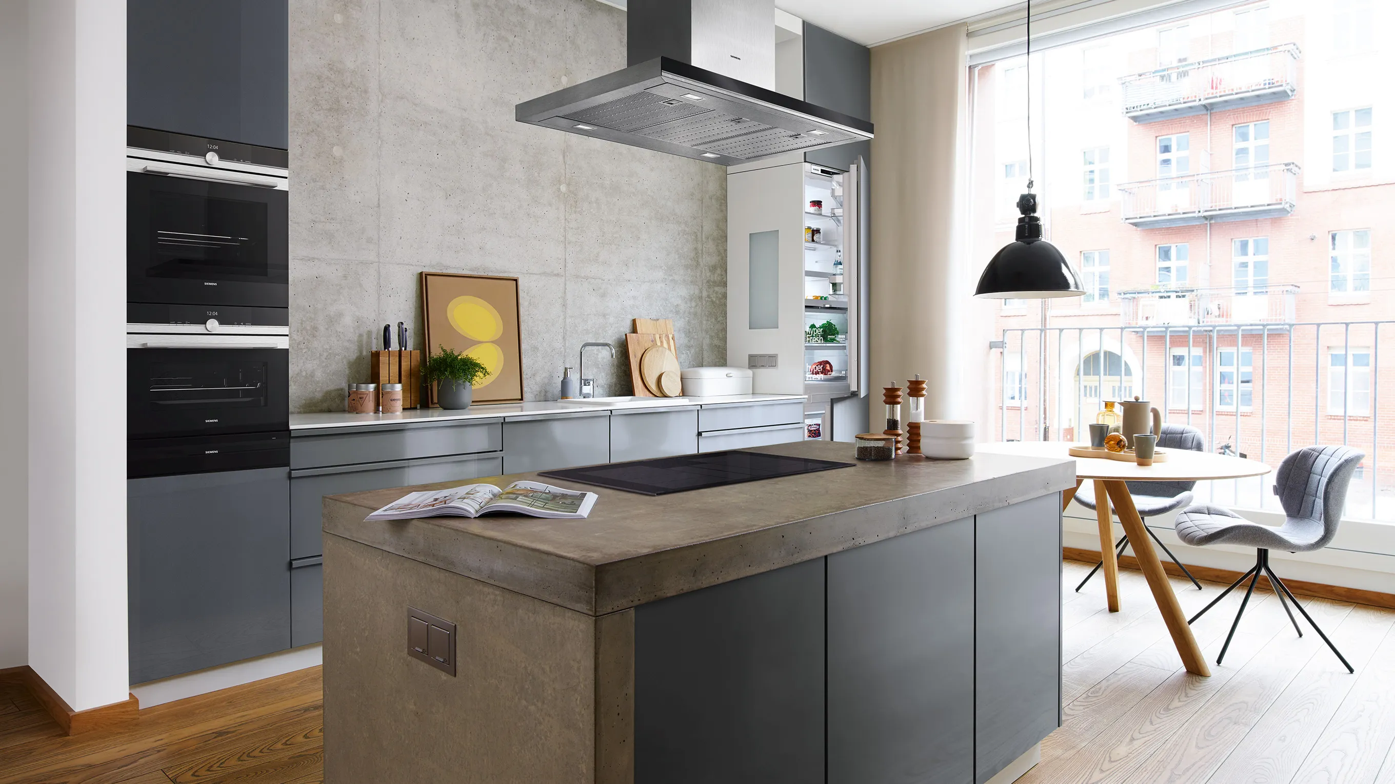 Contemporary kitchen with a concrete-look island, matte gray cabinets, built-in ovens and hood, and a bright dining nook by a large window.