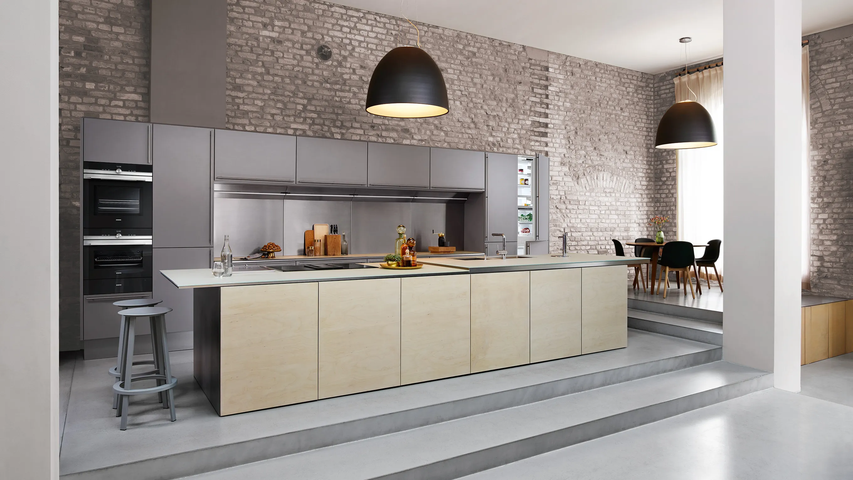 Modern open-plan kitchen with a large light-wood island, stainless built-in appliances, exposed brick wall, and raised dining area.